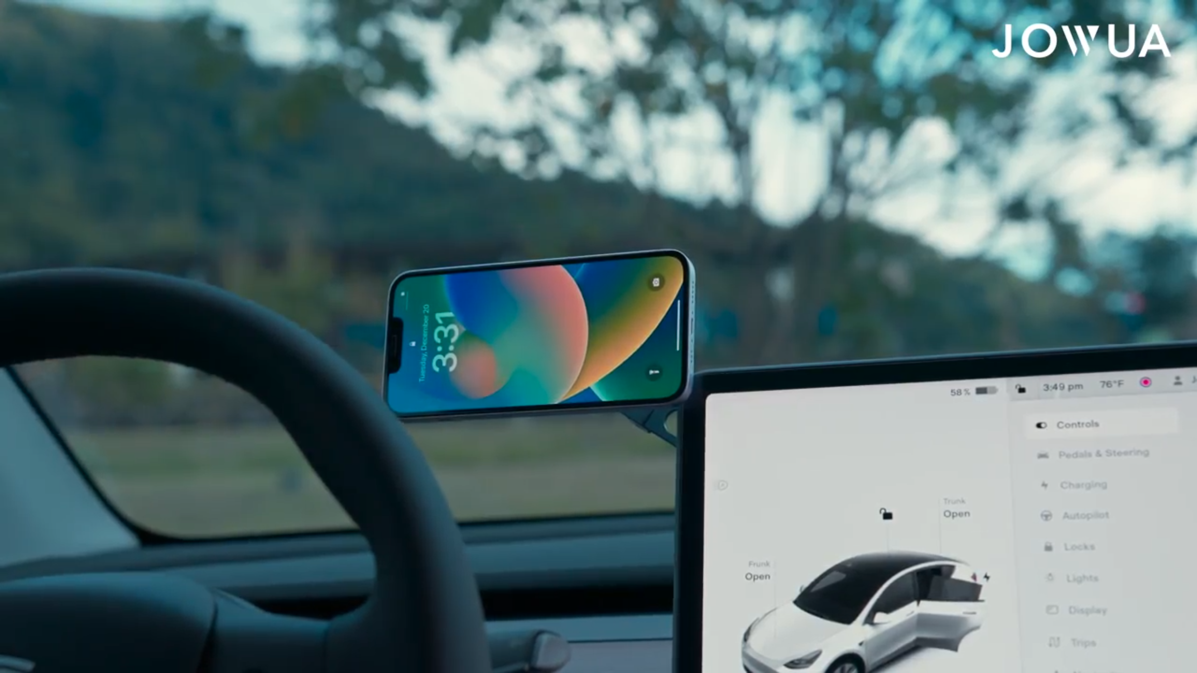 A smartphone on the JOWUA 6D Tesla Compatible Invisible Foldaway Car Mount Qi2 Wireless Charger sits by the steering wheel displaying the time, while a tablet shows car controls. Trees and a hill are visible through the windshield.