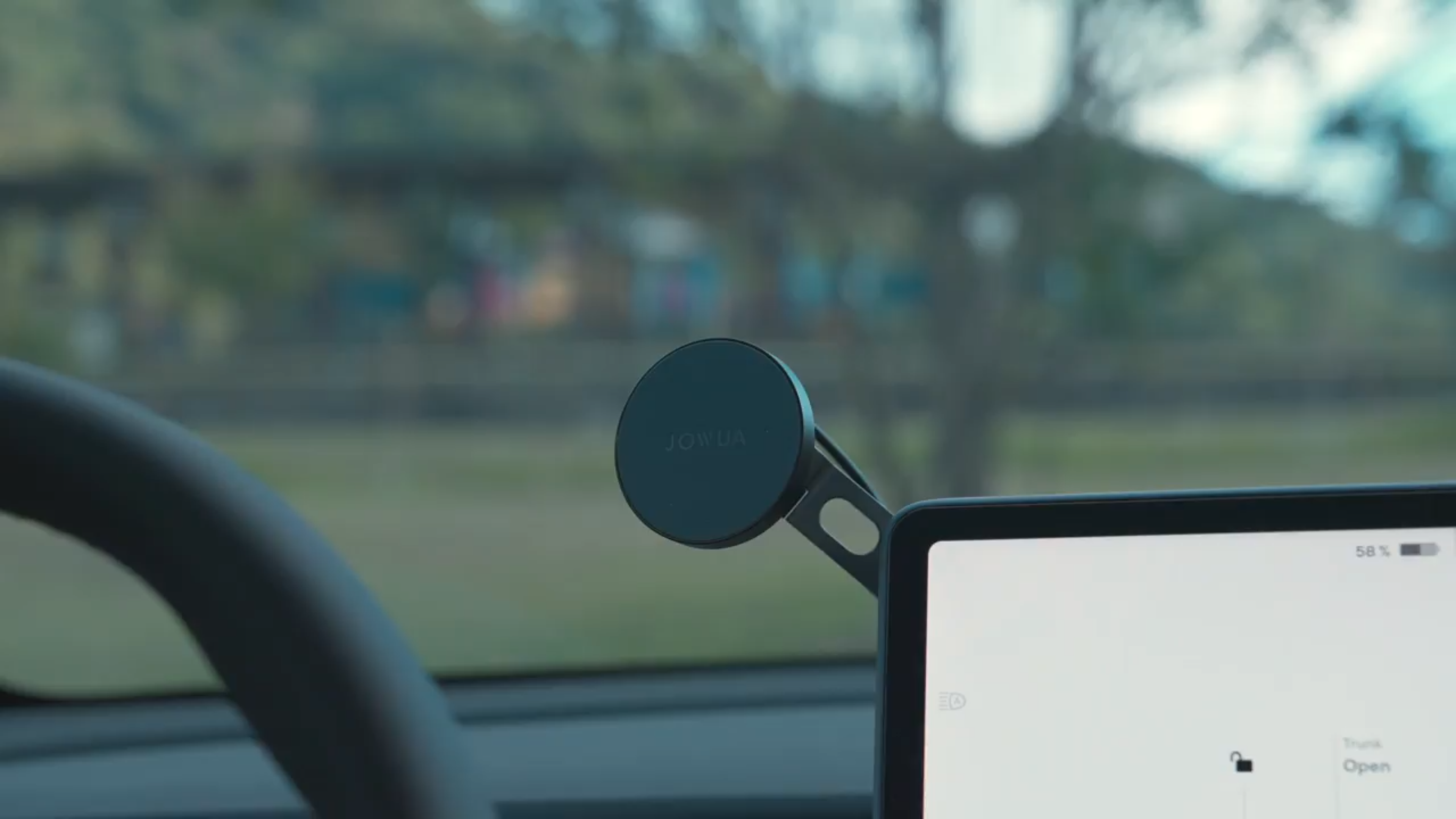 A close-up of a Tesla dashboard shows a circular JOWUA 6D Invisible Foldaway Car Mount Qi2 Wireless Charger (MagSafe) beside the digital display, with a blurred outdoor view through the windshield.