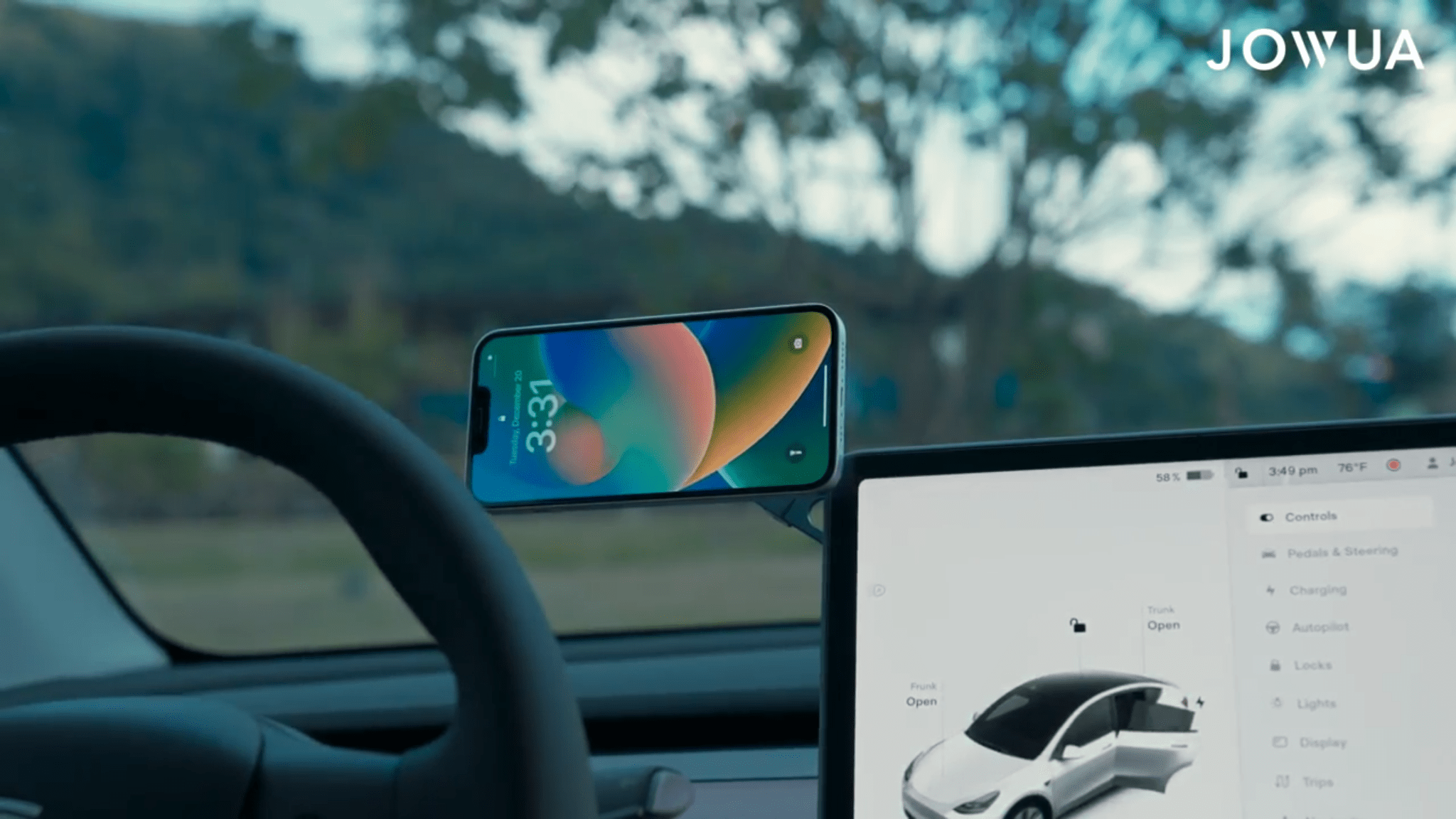 A smartphone on the JOWUA 6D Tesla Compatible Invisible Foldaway Car Mount Qi2 Wireless Charger sits by the steering wheel displaying the time, while a tablet shows car controls. Trees and a hill are visible through the windshield.