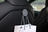 A round gray hook from the JOWUA Premium Interior Essentials Set – Model Y Juniper hangs on a Tesla Model Y headrest, holding a white shopping bag in the dim cabin, with a faint outdoor view through the window.