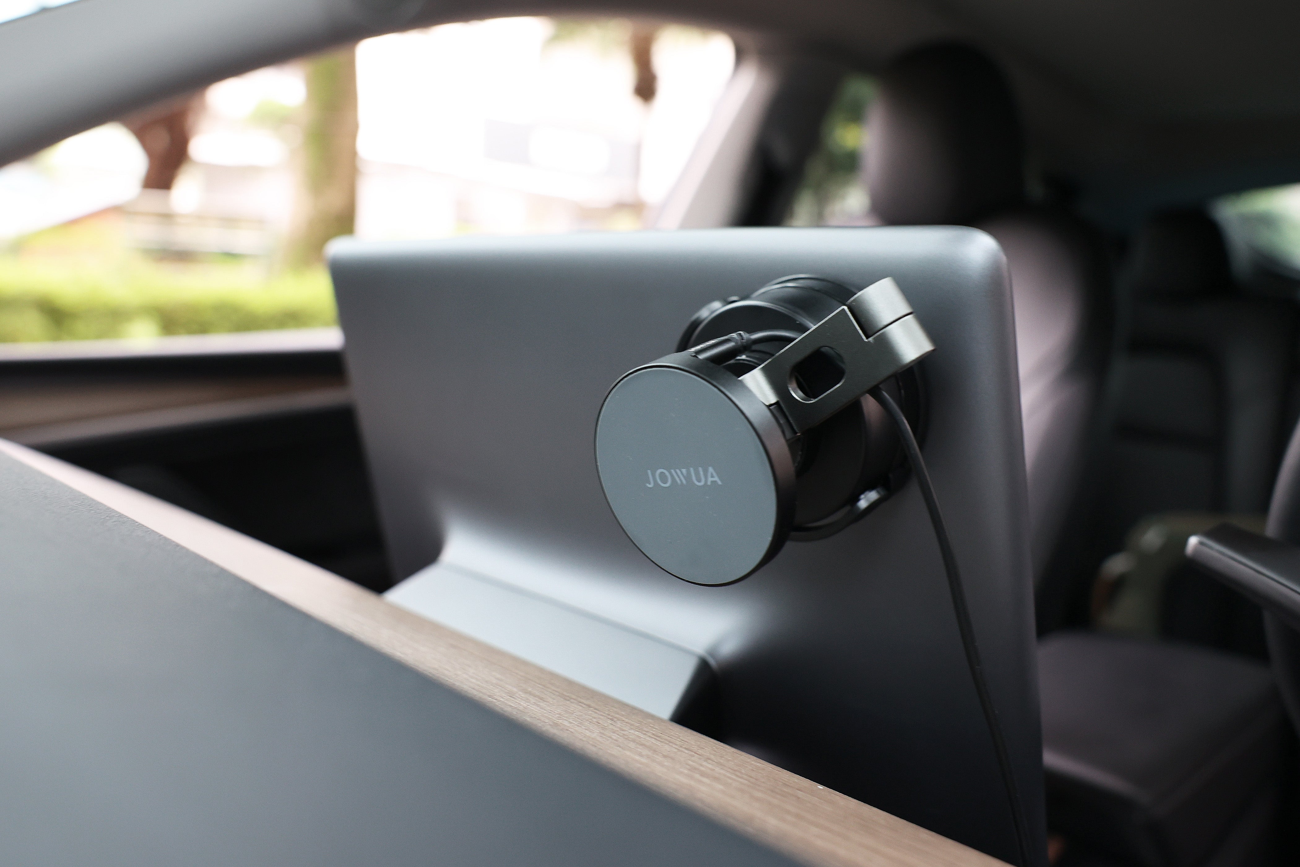 A "JOWUA" 6D Tesla Compatible Invisible Foldaway Car Mount Qi2 Wireless Charger for Tesla S/3/X/Y (MagSafe), featuring an adjustable metal arm, is secured to the back of a car seat headrest. The sleek wooden dashboard and rear seat inside the car enhance its elegant interior, while the view through the windows of blurred greenery indicates that the car is parked and ready for adventure.