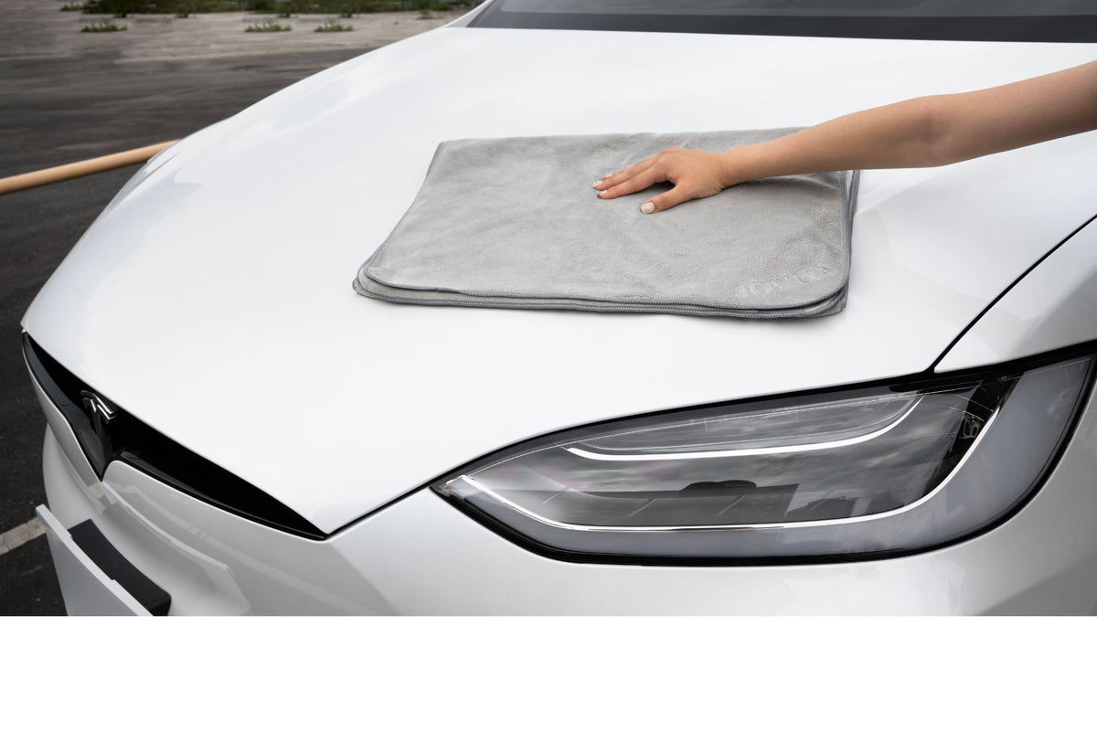 A person uses the JOWUA Under Seat Collapsible Organizer & Microfiber Cleaning Cloth Combo to clean the hood of a white Tesla with a gray microfiber cloth.
