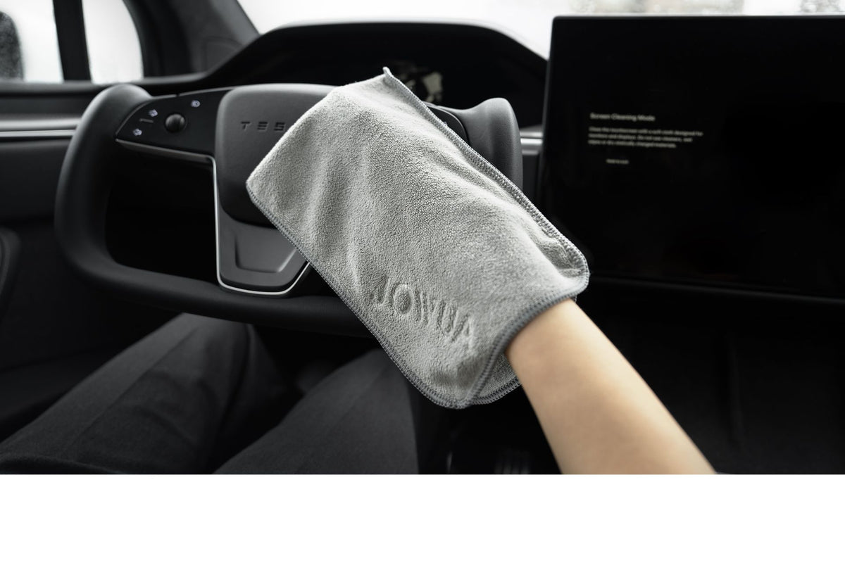 A person uses the JOWUA Microfiber Cleaning Cloth to wipe a Tesla steering wheel, with the JOWUA Under Seat Collapsible Organizer visible beneath the seat and the dashboard and touchscreen in view.