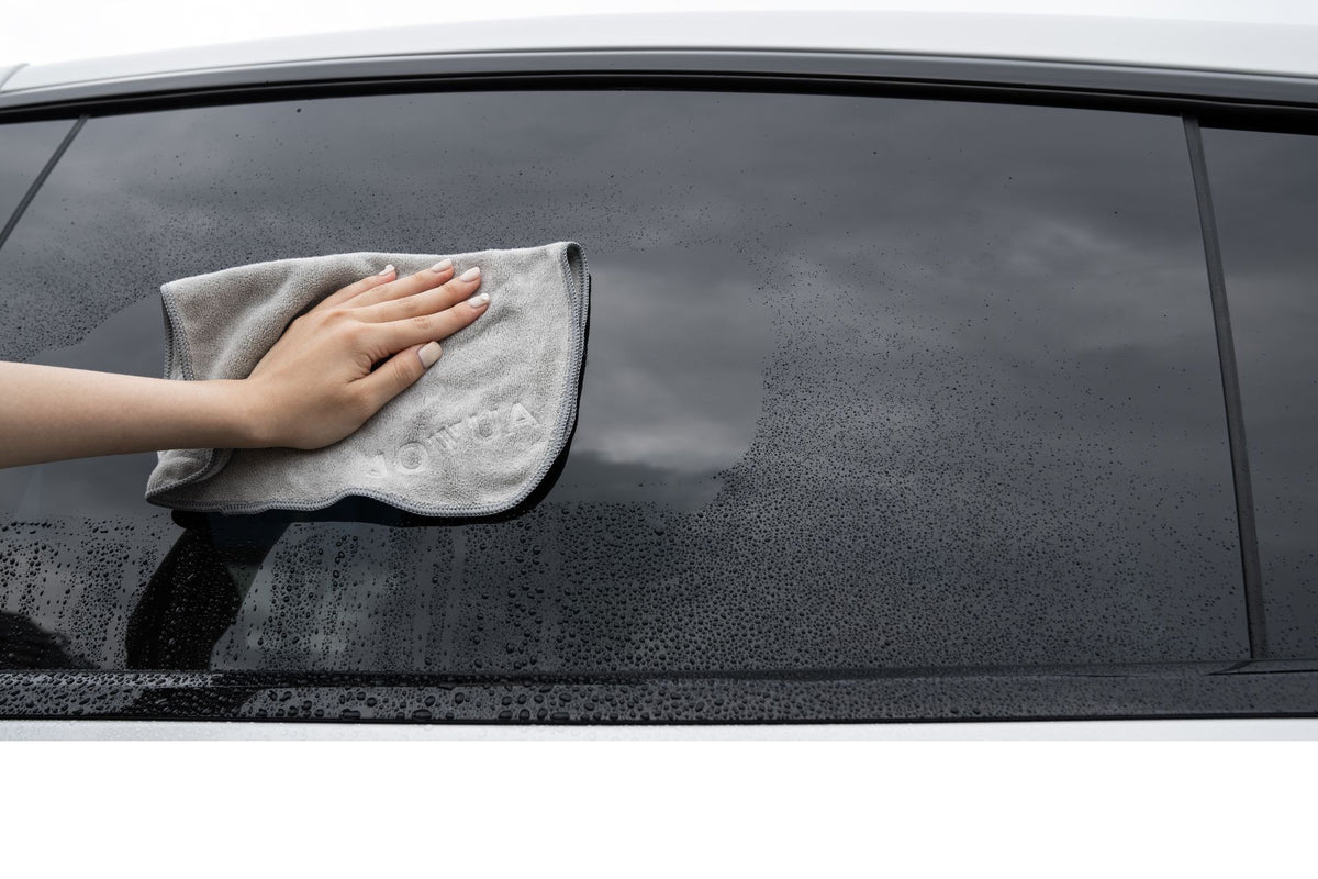 On a cloudy day, someone uses the JOWUA Under Seat Collapsible Organizer & Microfiber Cleaning Cloth Combo to wipe water droplets from a car window, holding the gray cloth against the glass after retrieving it from under the seat.