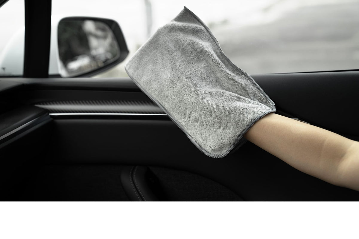 A hand uses the JOWUA Microfiber Cleaning Cloth from the Under Seat Collapsible Organizer & Microfiber Cleaning Cloth Combo to clean a car door near the window, with the side mirror and blurred exterior scenery visible outside.