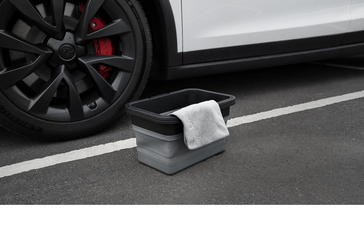 A JOWUA Under Seat Collapsible Organizer & Microfiber Cleaning Cloth Combo sits on asphalt near the wheel of a white car parked in a lot.