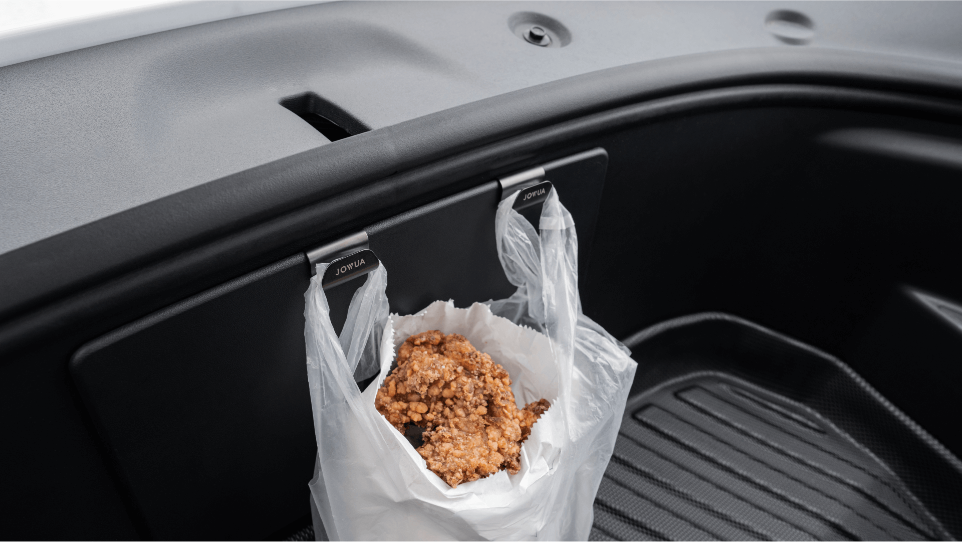 Inside the car, a white plastic bag with a brown, crumbly object hangs from a JOWUA Hook Bundle Model: 3 Highland 2024+ on the front trunk. The interior is dark and sleek with a visible rubber floor mat.