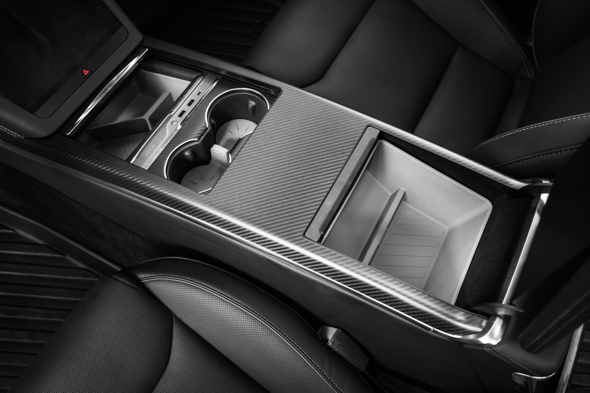 Black and white photo showcasing the Armrest Organizer for Tesla Model S/X by JOWUA. This product features cup holders and an open storage compartment, designed with a sleek, modern aesthetic crafted from premium hybrid material. The surrounding seats seem to be upholstered in leather.