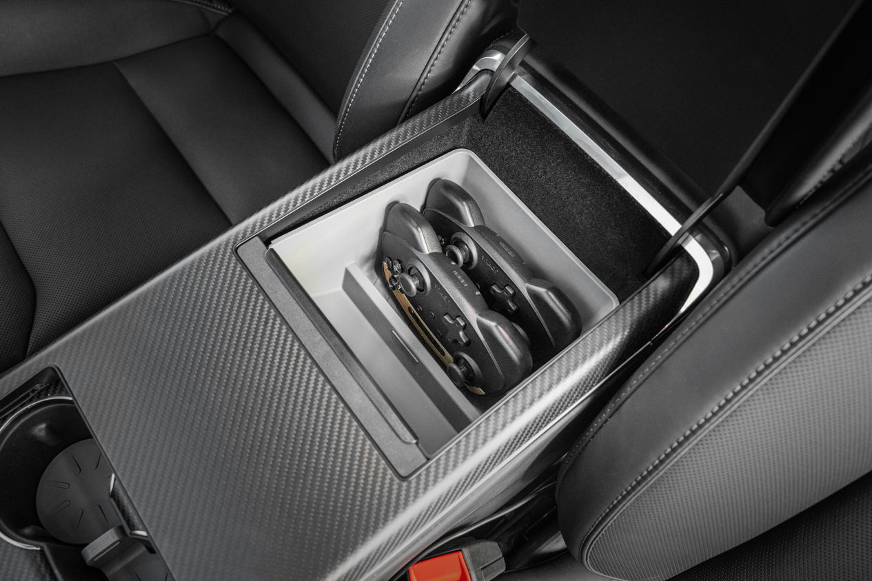 A car interior featuring the JOWUA Armrest Organizer for Tesla Model S/X, with an open center console crafted from premium hybrid material and a carbon fiber finish. The console houses two game controllers, while the surrounding seat and console materials are dark gray with accent stitching.