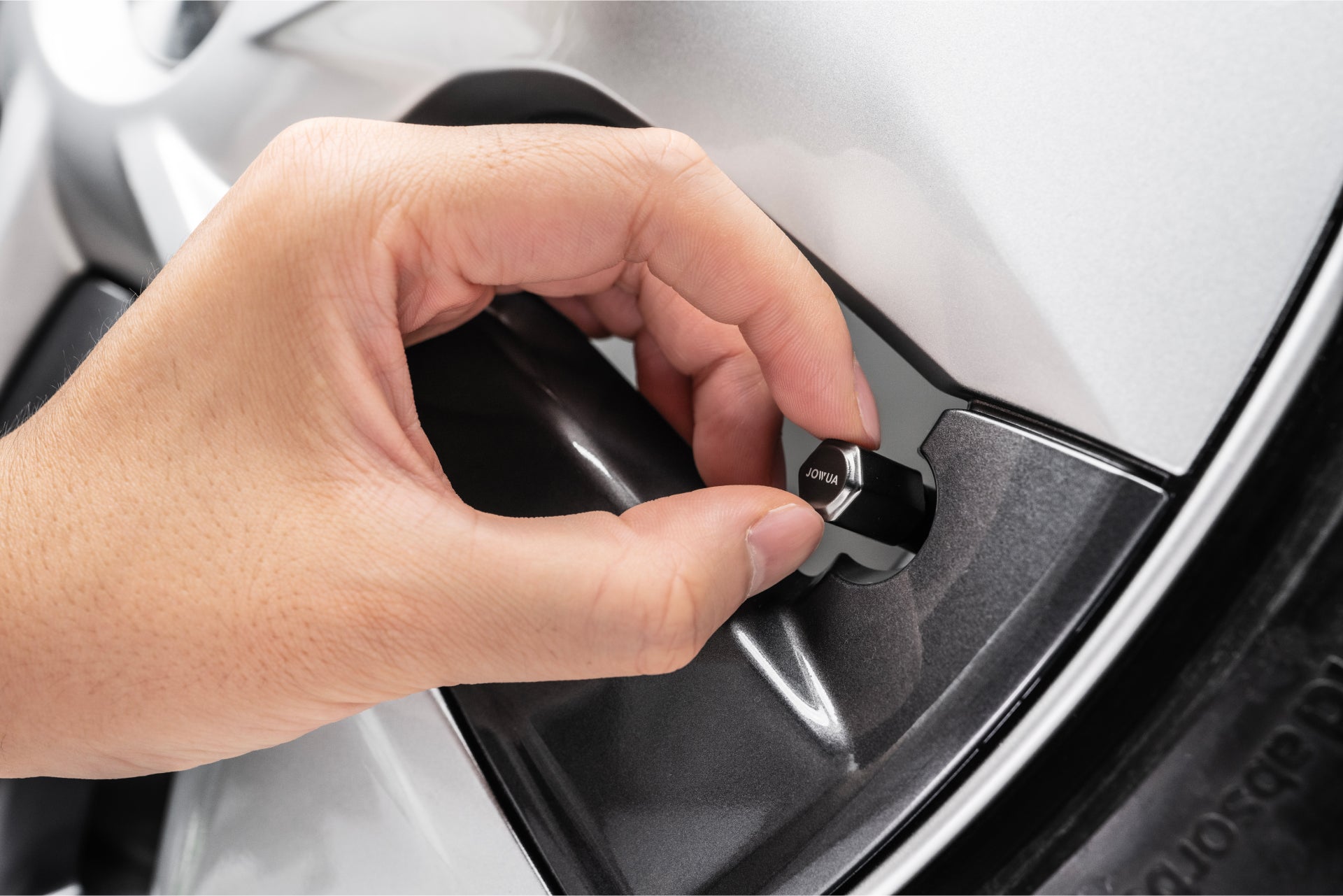 A hand is interacting with a JOWUA Tire Valve Cap, designed in a silver finish, similar in appearance to zinc alloy.