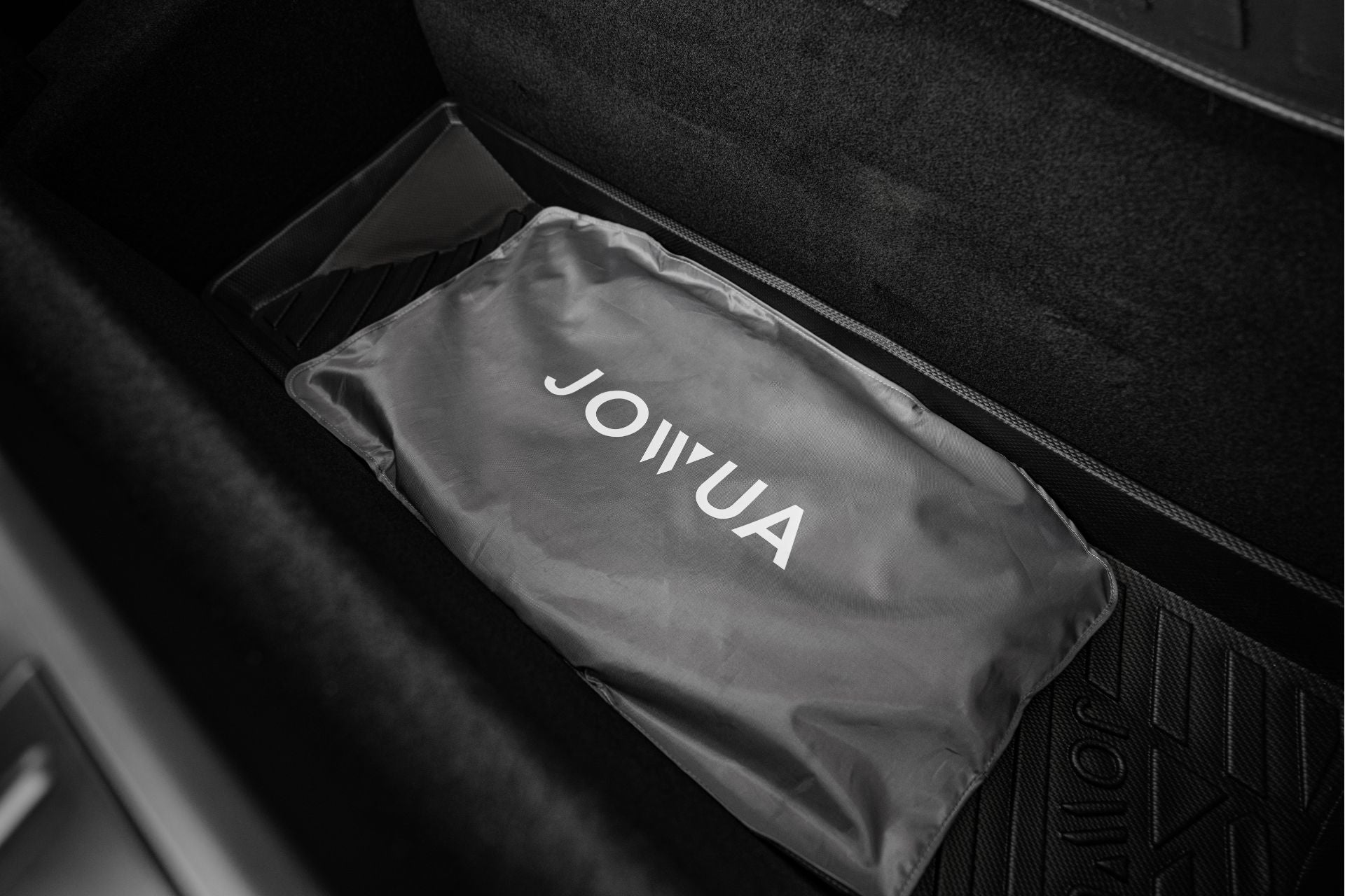 A black and gray "JOWUA" Glass Roof Sunshade- Separate Design (Model X) bag is neatly placed in the car's trunk, showcasing sunroof-inspired elegance. The dark-toned trunk area features textured floor mats, suggesting sophisticated UV ray protection.
