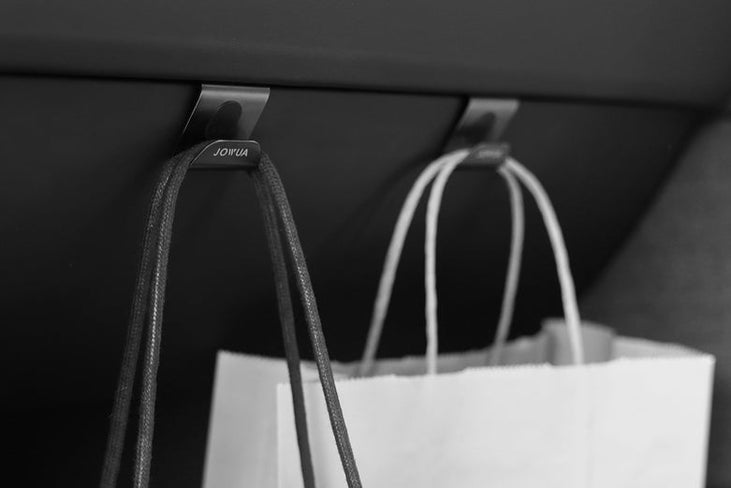 Close-up of two shopping bags on a glovebox hook under a car dashboard. The hooks, labeled "JOWUA," belong to the Hook Bundle Model: 3 Highland 2024+. One bag has black handles, the other white. The scene is in black and white.