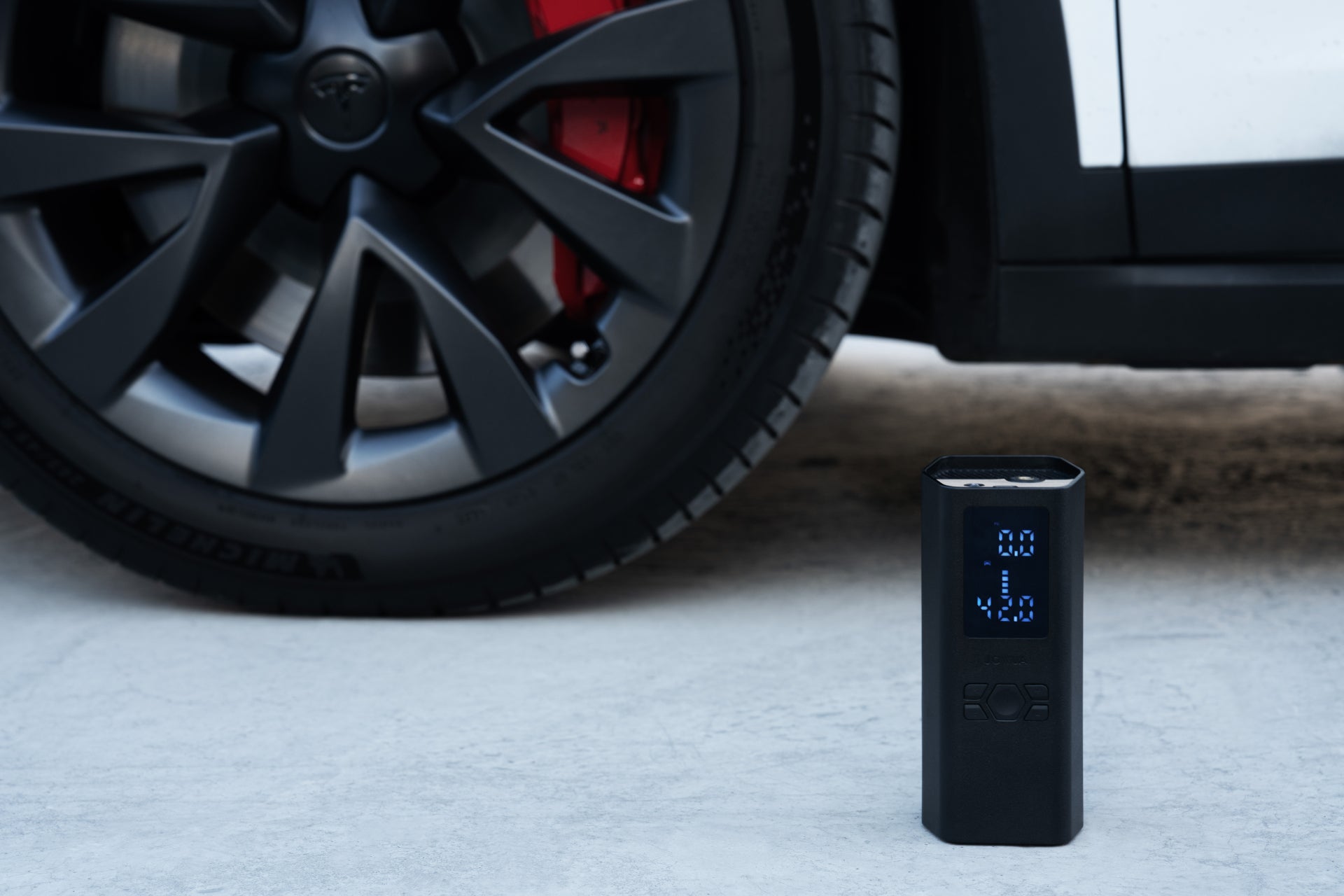 The JOWUA Portable Air Compressor + Charging Solution Kit, featuring a blue LED display, stands upright before a black alloy wheel—ideal for inflating Tesla tires with fast charging convenience.