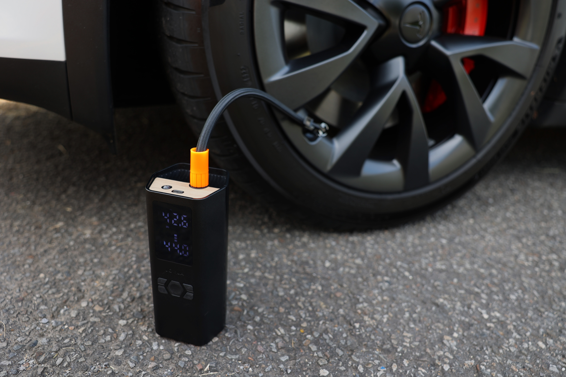 The JOWUA Portable Air Compressor + Charging Solution Kit is inflating Tesla tires on a paved surface, displaying digital readings on its screen and connecting securely to the tire valve with its attached hose.