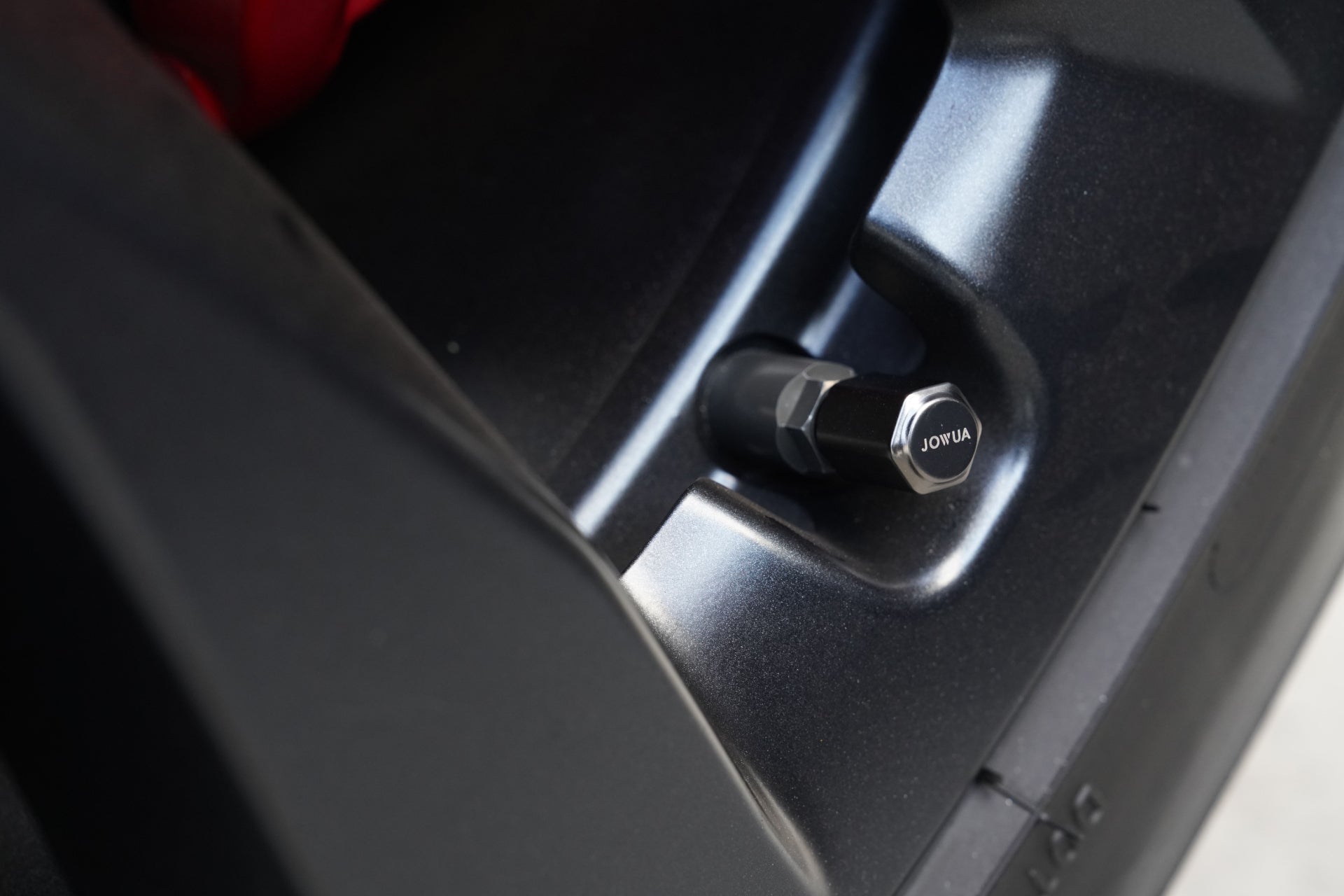 Close-up of a tire's valve stem cap on a vehicle rim. The metallic cap, inscribed with "JOWUA," complements the glossy black rim and its matte surroundings, pairing perfectly with Tesla accessories for those seeking elegance and high-speed charging convenience with the JOWUA 100W Portable Air Compressor + Charging Solution Kit.