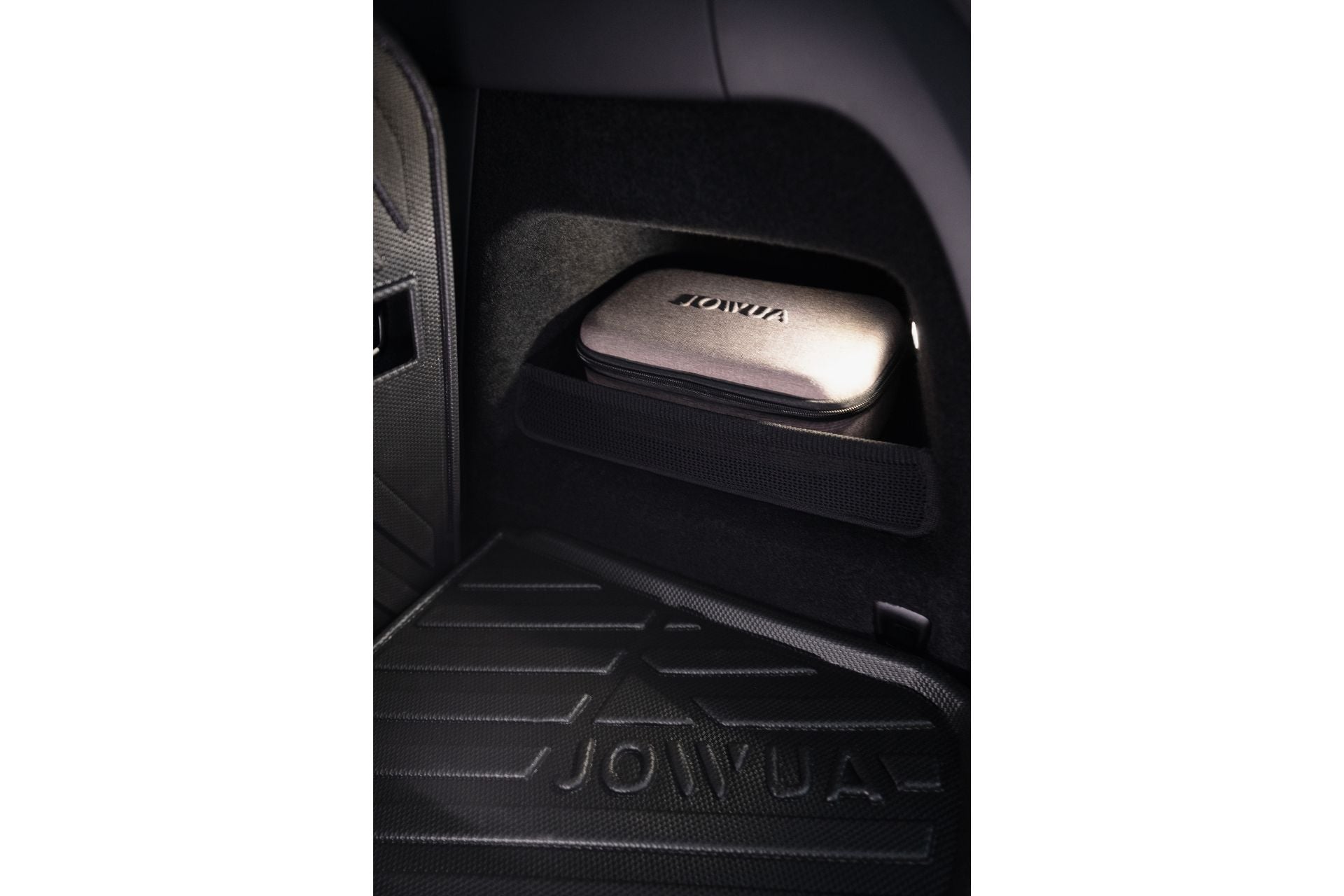 A compact 100W Portable Air Compressor by JOWUA sits neatly on an EV shelf with a small securing net. The car floor mat displays the "Jowua" logo below. Dimly lit, this gray device ensures fast inflation when needed.