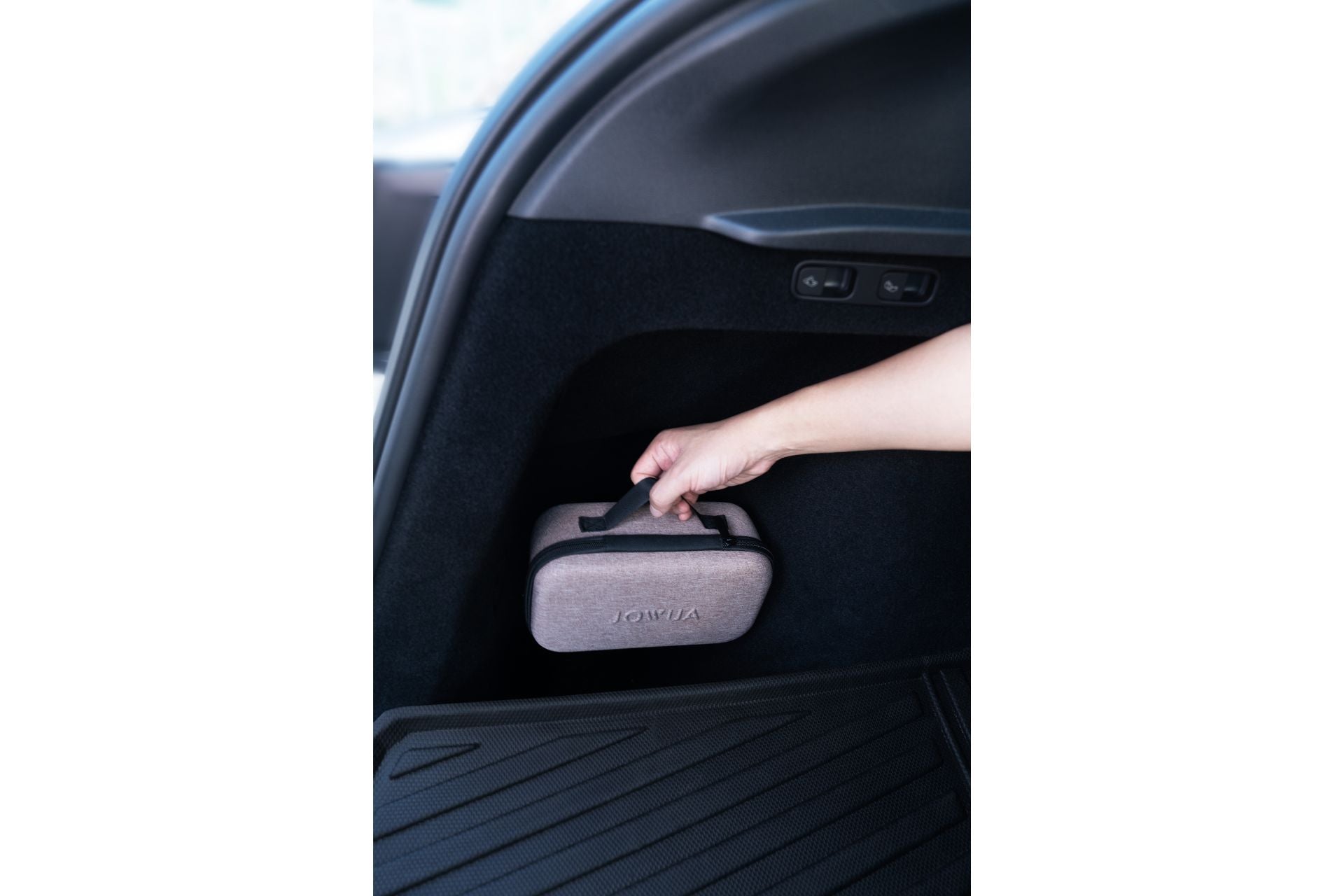 A person places a gray fabric case with a handle containing JOWUA's 100W Portable Air Compressor + Charging Solution Kit inside a car trunk. This ideal spot for Tesla accessories features a black textured interior with buttons above the case and a rubber mat lining the floor.