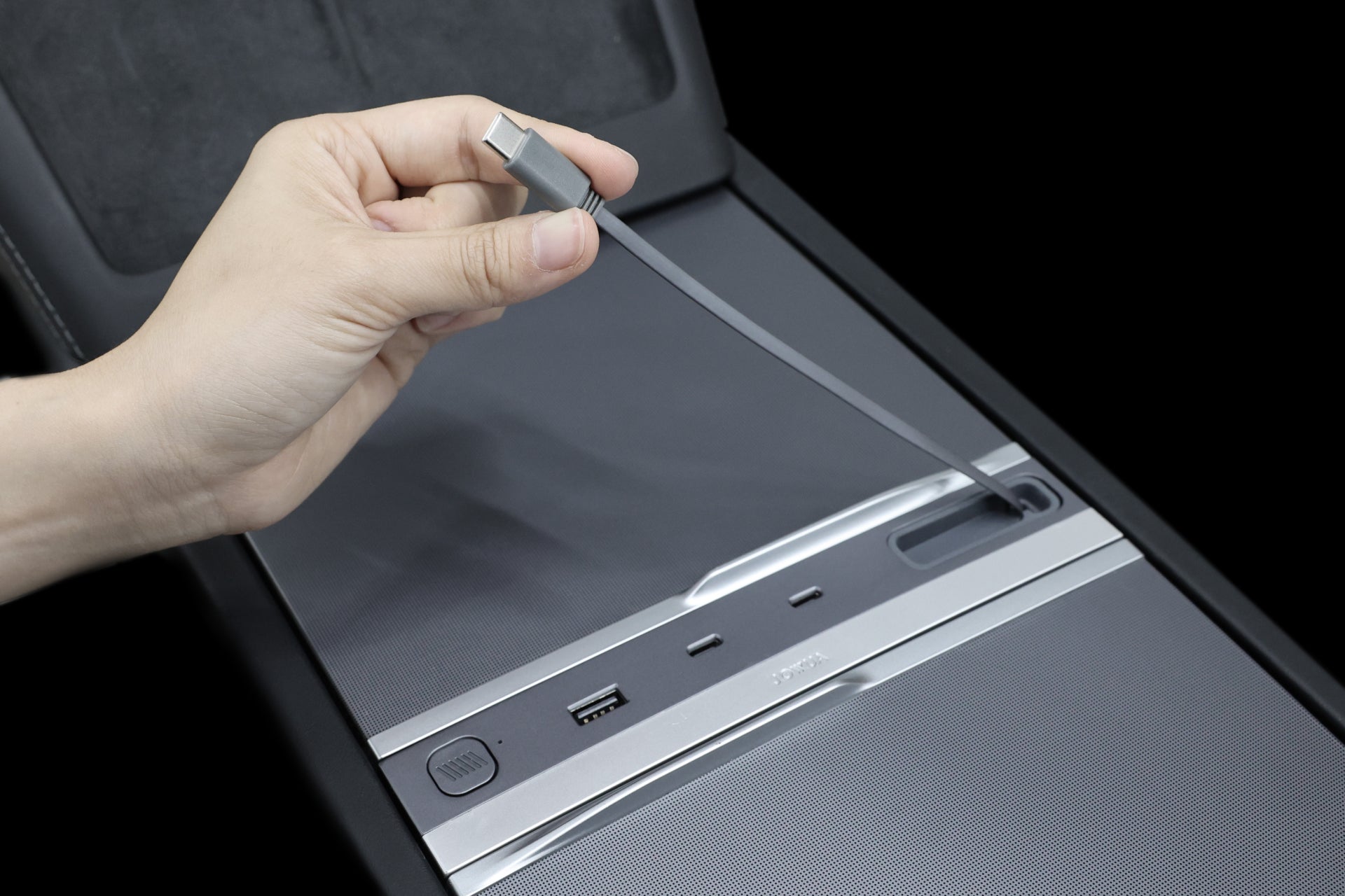 A hand holds a charging cable with a USB-C connector above JOWUA's sleek 130W LED Hub for Tesla Model Y Juniper 2025+ / Model 3 2024+, featuring a USB port, an SD slot, and control buttons on its elegant surface, providing both charging and storage solutions.