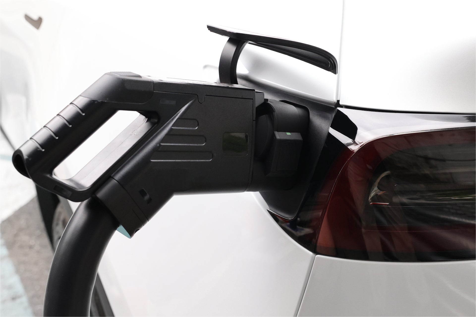 Close-up of a white electric car utilizing the JOWUA CCS1 to CCS2 Adapter for charging at a supercharging station. A black charging cable, attached with the adapter, is connected to the vehicle's charging port near the rear taillight. The car is parked in a designated spot for DC fast charging.