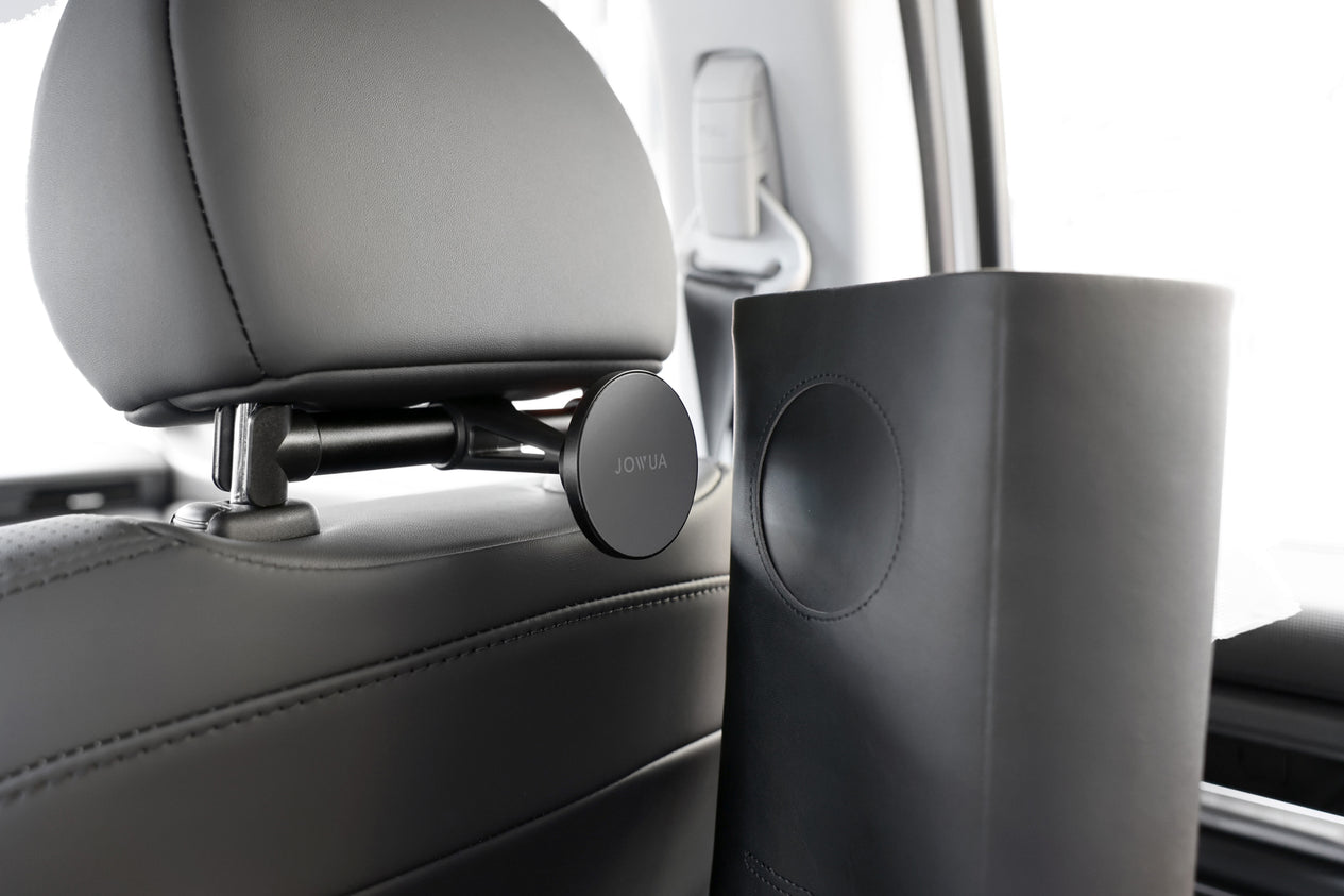 A car interior displaying a black leather-covered headrest featuring the JOWUA Universal MagSafe Car Seat Holder mounted on its back. A cylindrical leather bag is partially visible on the rear seat. The well-lit scene emphasizes the sleek, modern design of these car accessories with retractable features.