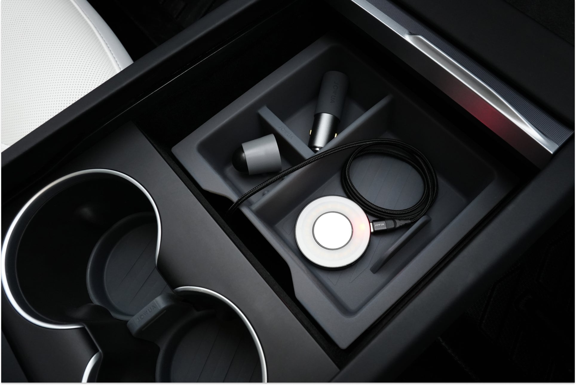 The JOWUA Center Console Set for the Tesla Model 3 Highland 2024+ opens up to reveal an organized storage space, featuring two cup holders on the left, each fitted with a cup coaster. On the right side, there is a silicone organizer that holds chargers and a cable efficiently. The interior design showcases a sleek and modern matte finish.