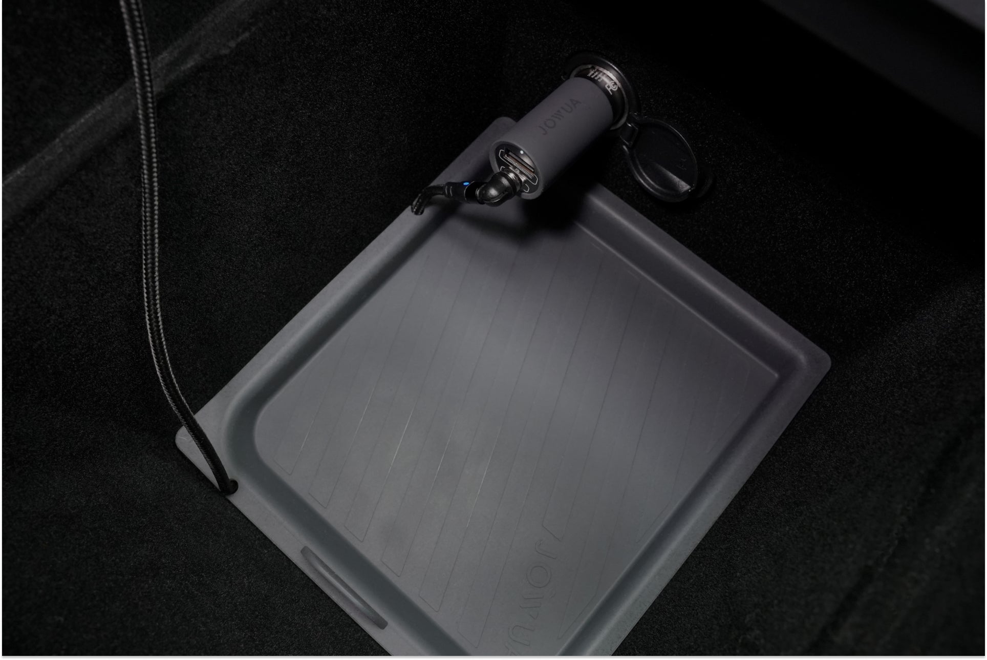 In the car's trunk space, a rectangular storage tray made from premium silicone showcases a portable air pump or vacuum cleaner. The sleek and modern dark grey design provides an elegant contrast against the black carpeted background, highlighting the LED Center Console Combo by JOWUA for the Tesla Reengineered Model 3 Highland (2024+).