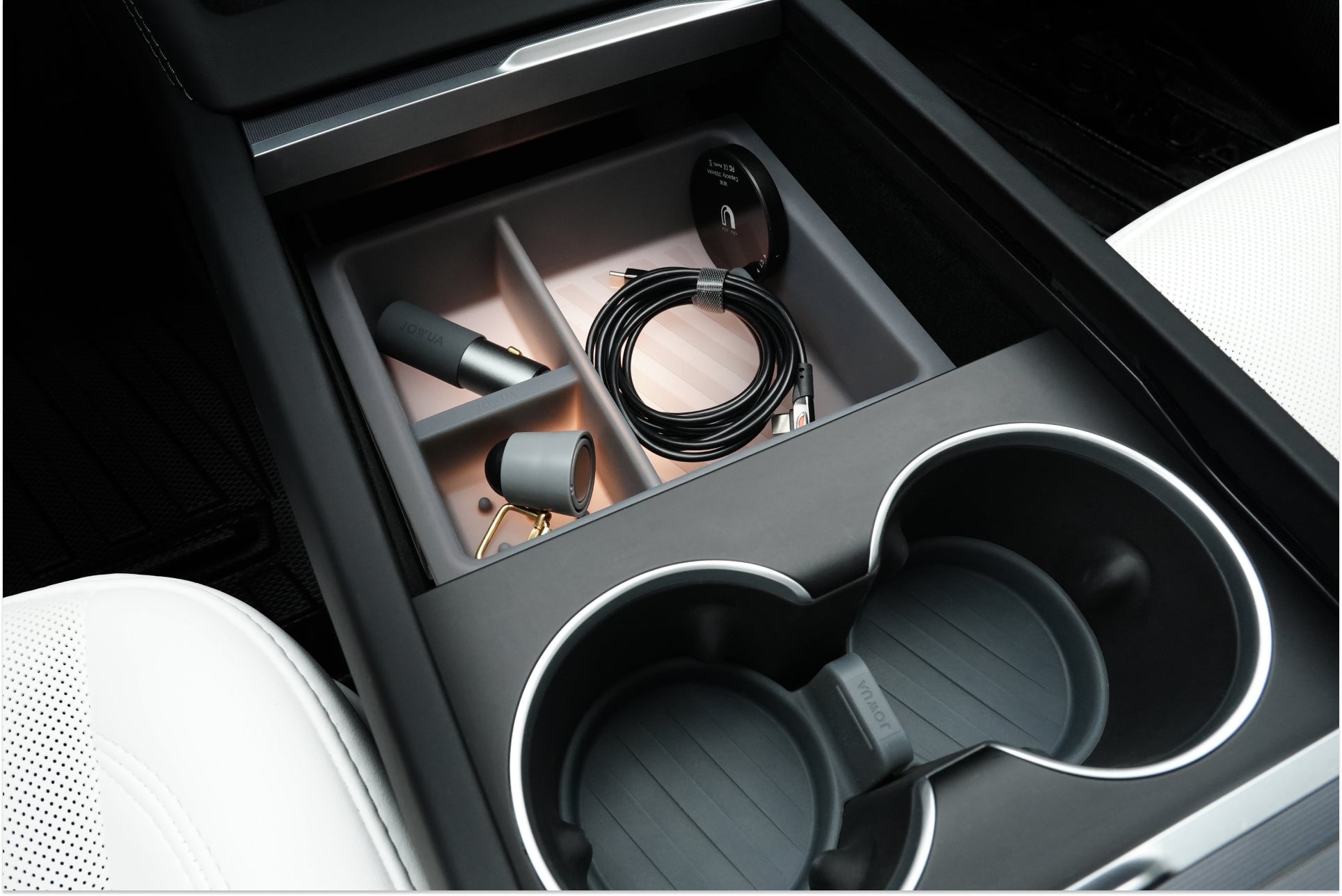 The JOWUA Center Console Set for the Tesla Model 3 Highland (2024+) features a sleek design complete with dual cup holders and a silicone organizer. Inside, it includes a ring-shaped device, a coiled cable, and two gray cylindrical objects, all harmoniously set against the backdrop of partially visible white seats.