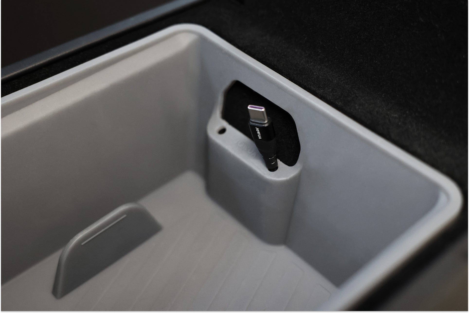 A close-up of the JOWUA Premium Interior Essentials Set – Model Y Juniper in gray, featuring a center console compartment with a built-in port holding a black charging cable with a visible USB-C or Lightning connector.