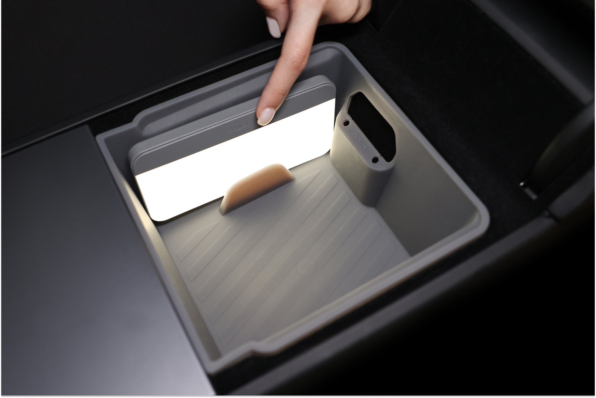 A hand points to a bright rectangular panel inside a ridged gray storage compartment, showcasing the JOWUA Premium Interior Essentials Set for Tesla Model Y Juniper as light illuminates the compartment’s interior.