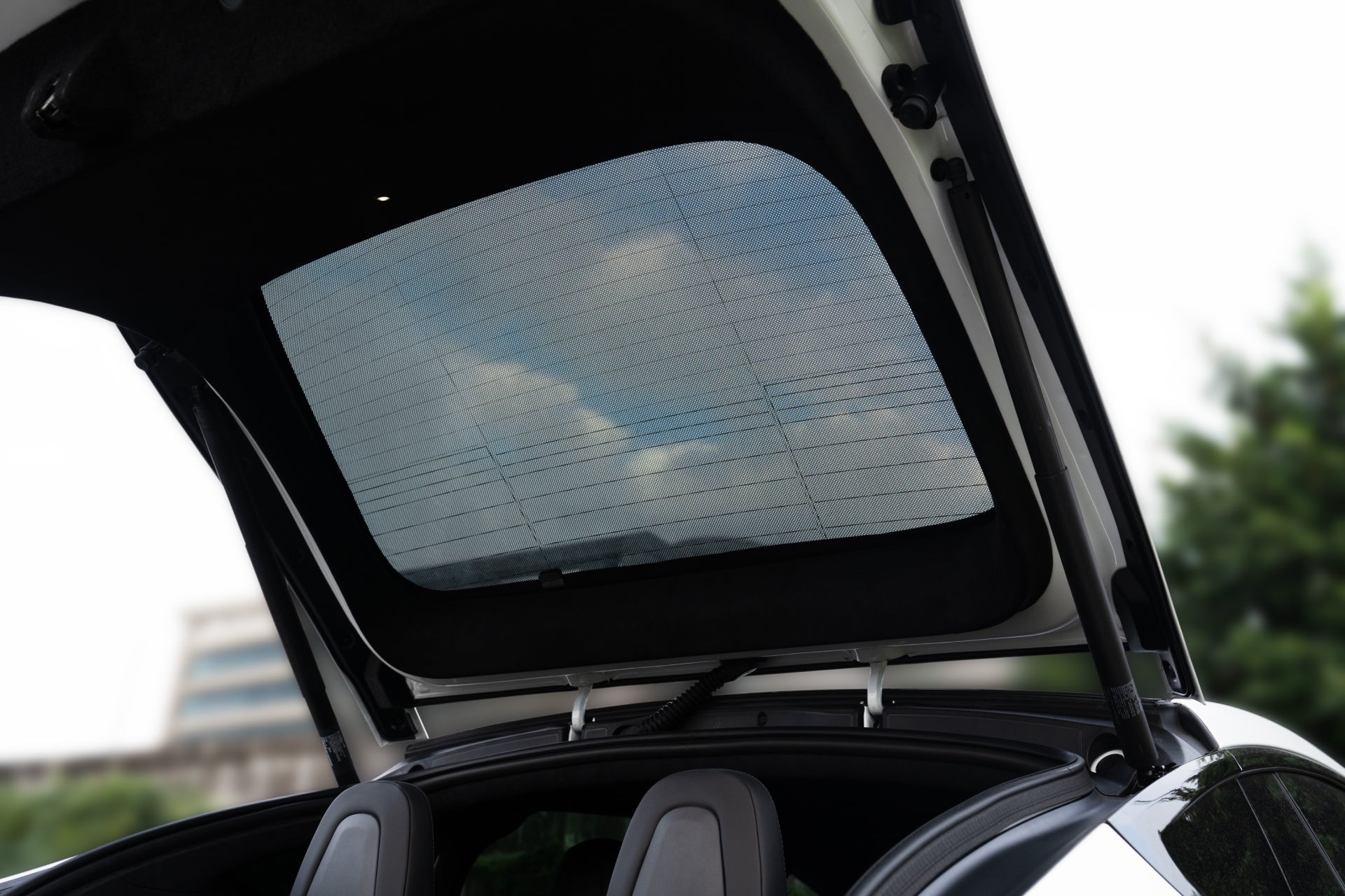 The image shows the open rear hatch of a car, with the rear windshield visible against a partly cloudy sky. A JOWUA Rear Lift Gate + Triangular Window Sunshade for Model X, crafted from high-density woven fabric, lines the interior roofline, offering shade over the two headrests of the backseat.