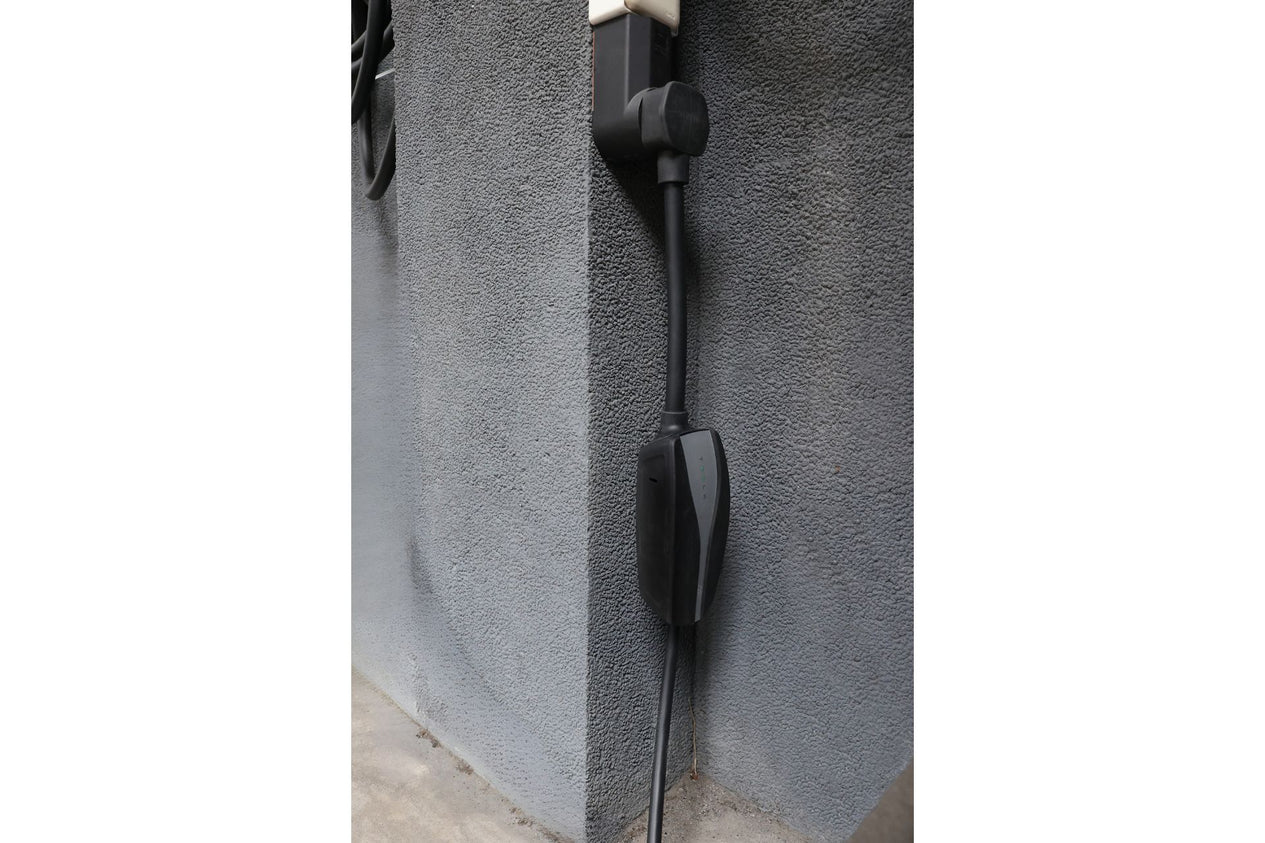 A JOWUA NEMA 14-50 Mobile Connector Adapter, designed for Tesla compatibility and featuring superior durability, is plugged into a wall outlet with a textured grey finish. The robust black charging cable hangs vertically against the wall, extending towards the ground.