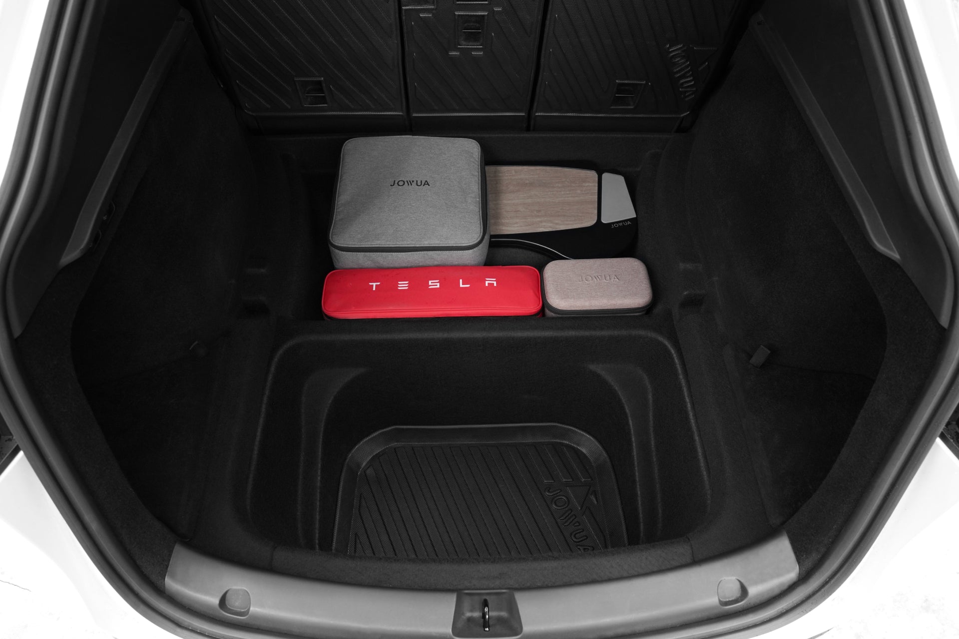 The open trunk of a Tesla Model Y showcases the JOWUA Model Y EasyShield Waterproof Trunk Liner + Rear Seats Back Cover Bundle, with several organized storage bags, including a red Tesla-branded case and two gray pouches neatly arranged.