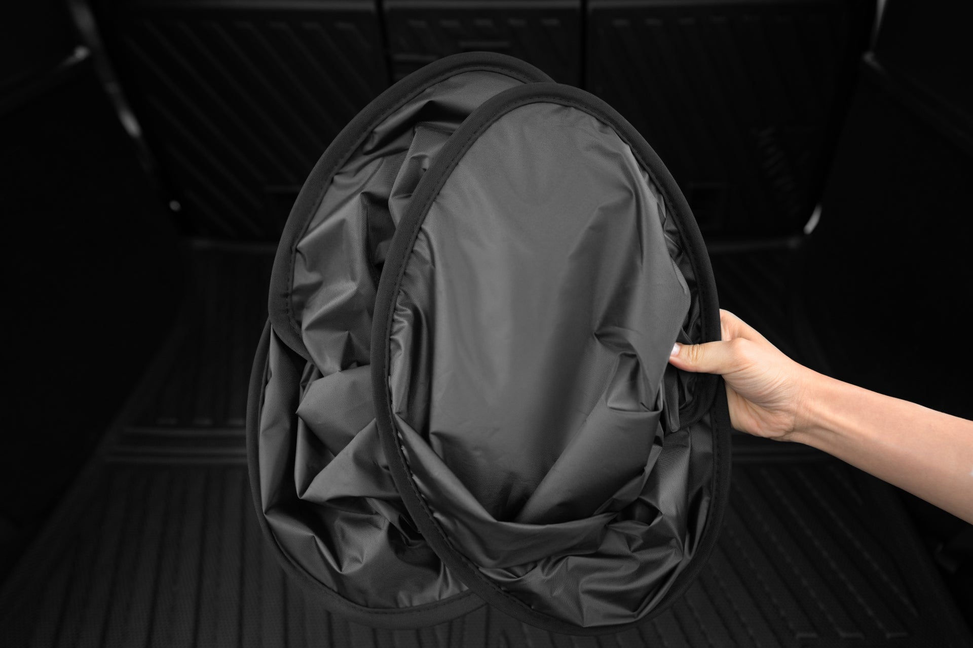 A hand holding two black foldable JOWUA Model Y Ultimate Sunshade Bundles in the back of a vehicle with a dark interior features Dark Matter mesh fabric, offering superior UV protection.
