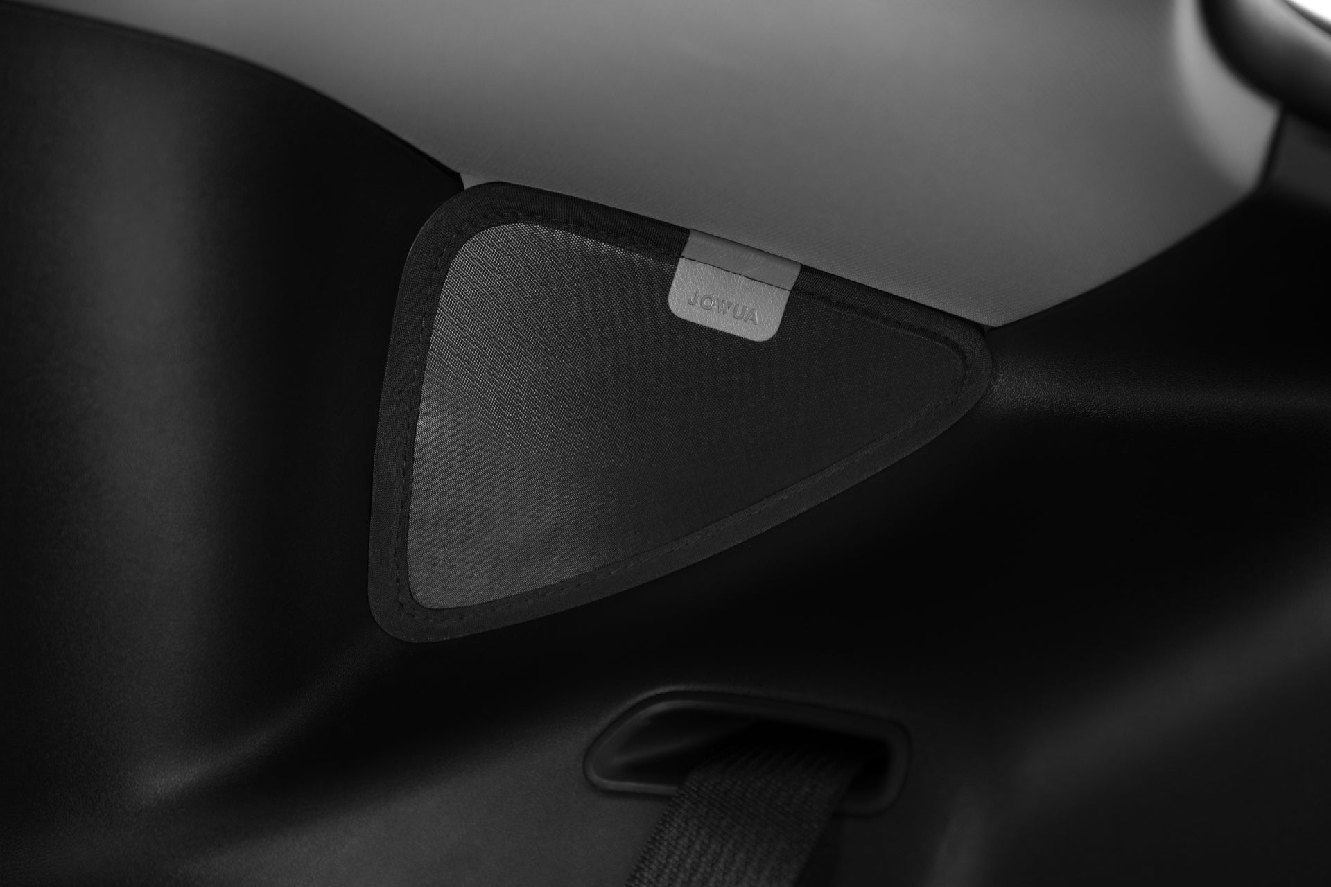 Close-up of the interior of a black car, showcasing a triangular speaker cover adorned with a small, rectangular tag bearing the brand name JOWUA. The cover is surrounded by textured material that suggests the sleek elegance characteristic of the Tesla Model Y. Below, you can see part of a seatbelt crafted from Dark Matter mesh fabric, engineered for UV protection as part of the Model Y Ultimate Sunshade Bundle.
