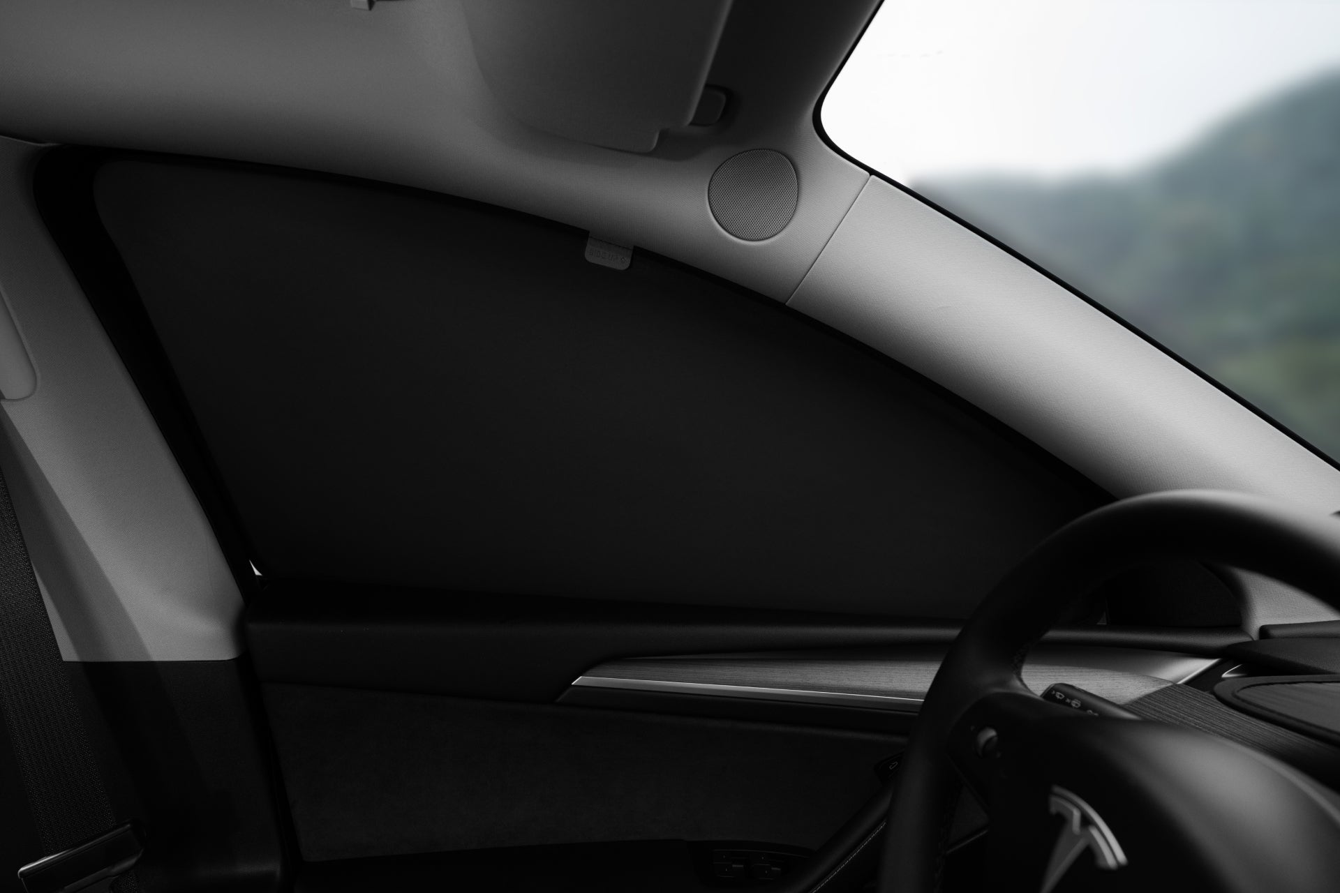 View from inside a car showcasing the driver's side dashboard and steering wheel, complemented by the JOWUA Model Y Ultimate Sunshade Bundle offering UV protection. The window is tinted black with Dark Matter mesh fabric, partially displaying the side mirror and interior features against a blurred landscape backdrop.
