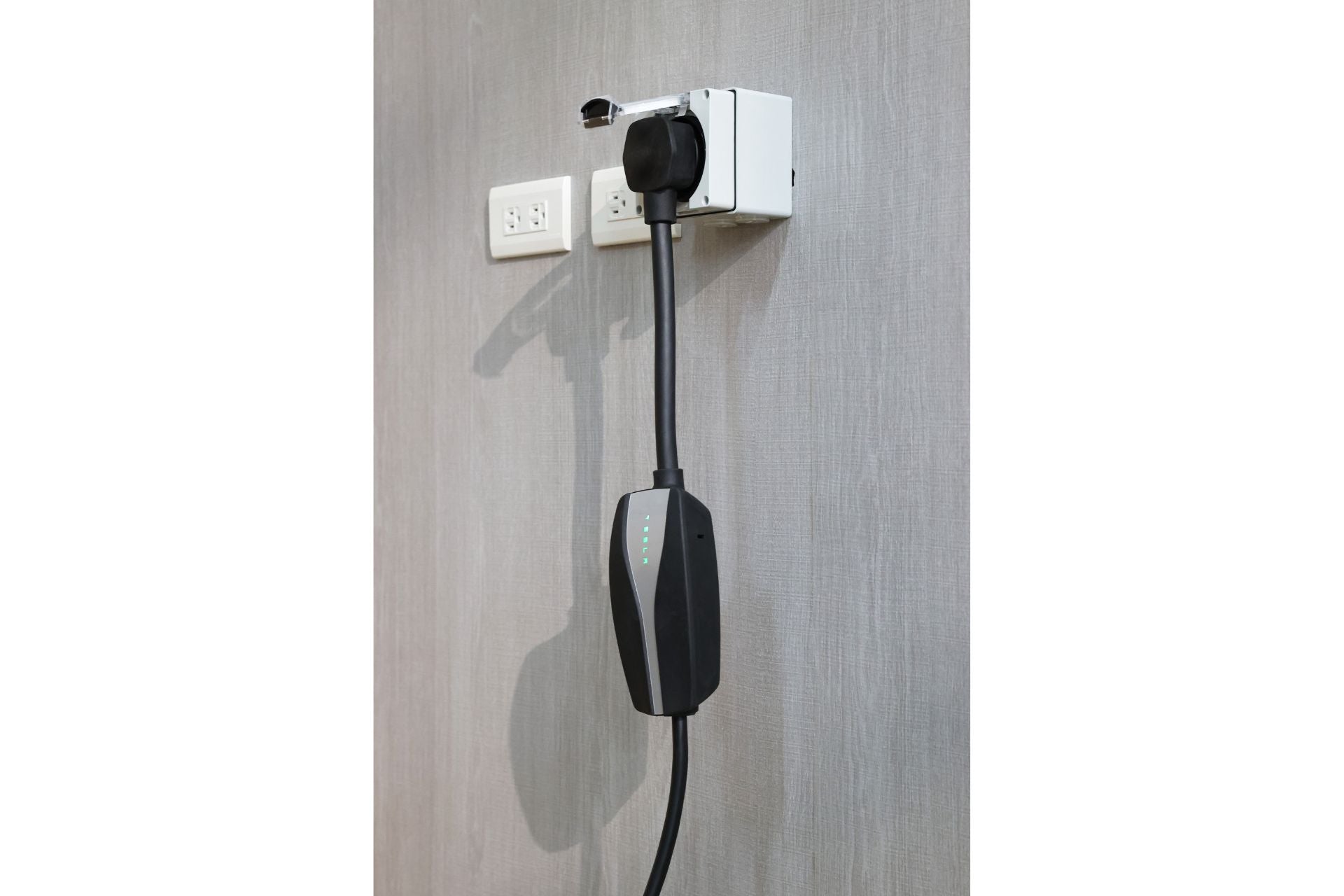 The JOWUA NEMA 14-50 Mobile Connector Adapter is shown in a wall-mounted electric vehicle charging station setup, featuring a black and gray charger unit connected via this Tesla-compatible adapter. A gray textured wall serves as the backdrop, featuring two standard electrical outlets that assure excellent cable durability.