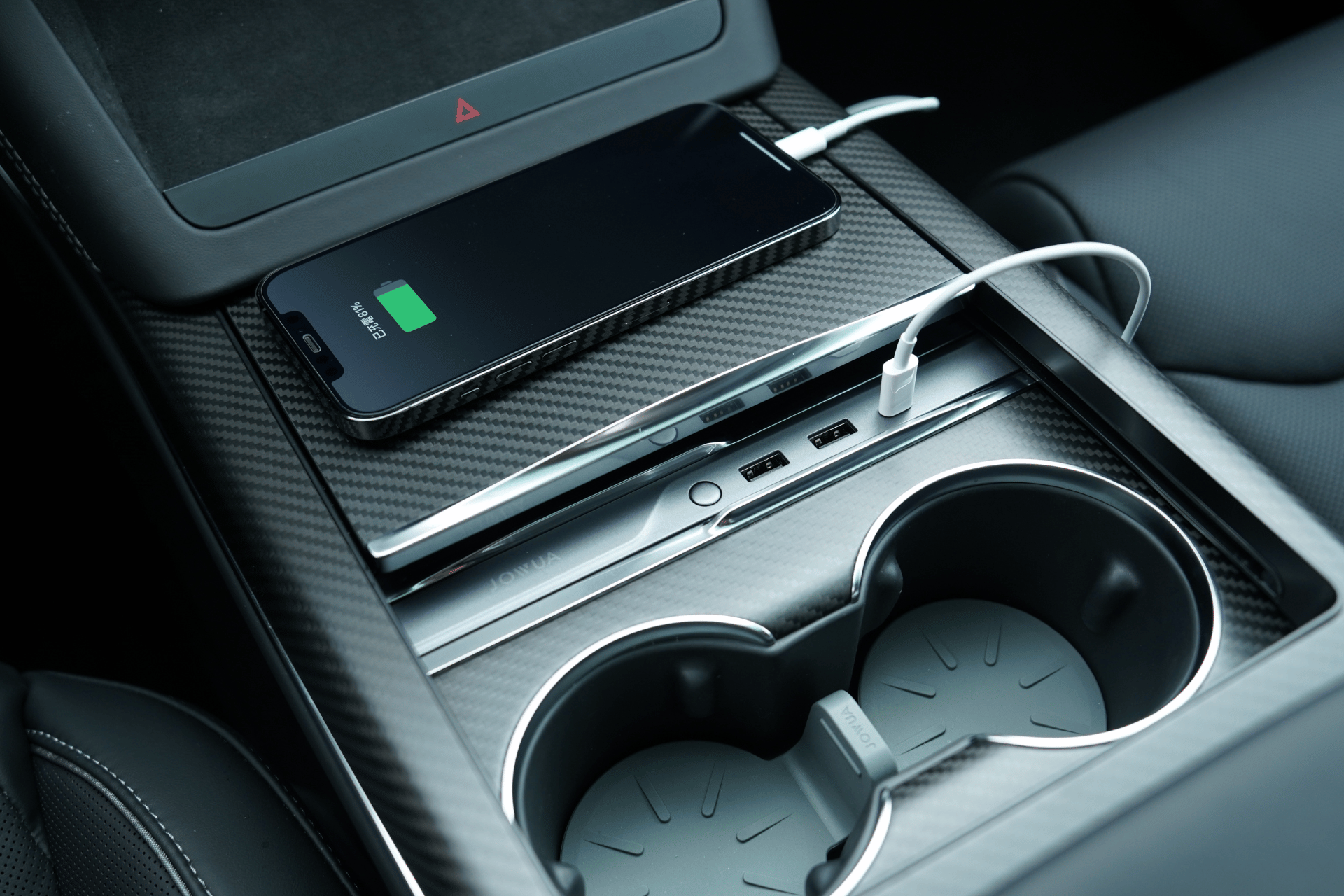 The JOWUA Hub Bar Mini for Tesla Model S/X charges your smartphone via cable on the center console, next to cup holders and multiple USB ports, with a carbon fiber finish enhancing the look.