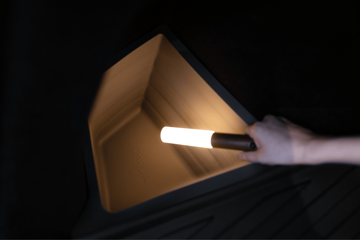 A hand holds a cylindrical flashlight, lighting up the corner of a dark, angular JOWUA Trunk Storage Bin Bundle for Model Y (2018-2024) with textured sides.