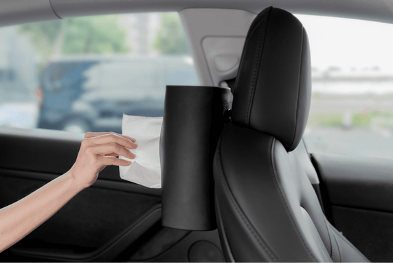 A hand reaches for a tissue from a cylindrical holder attached to JOWUA's Hook Bundle Model: 3 Highland 2024+ on the back of a car seat. The black leather interior contrasts with the blurred outdoor scenery through the window.