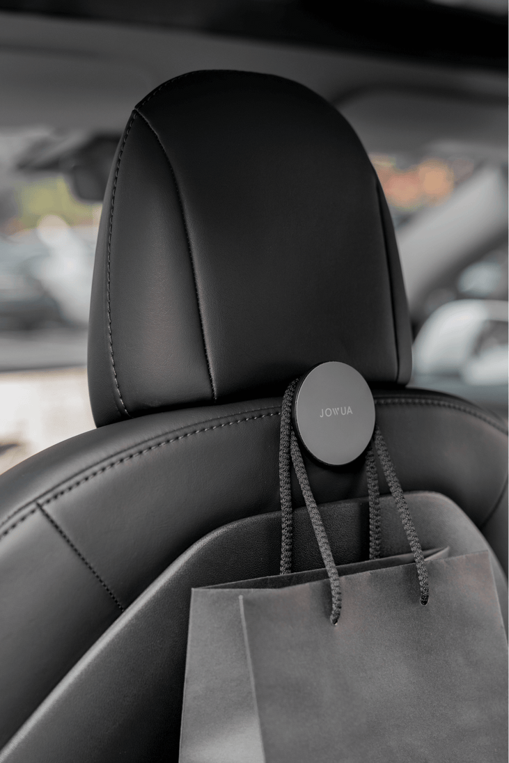 A black car seat displays a JOWUA-branded Hook Bundle for the Model 3 Highland 2024+, similar to a Glovebox Hook, attached to the headrest. A gray shopping bag with black handles hangs effortlessly, with softly blurred parked cars in the background.