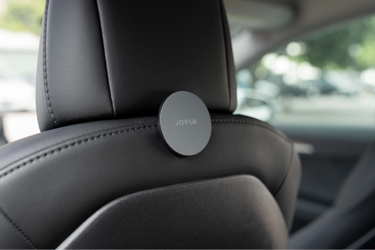 Close-up of a black leather car seat with a JOWUA Hook Bundle Model 3 Highland 2024+ mounted on the back of the headrest. The softly blurred interior reveals hints of greenery outside through the window.