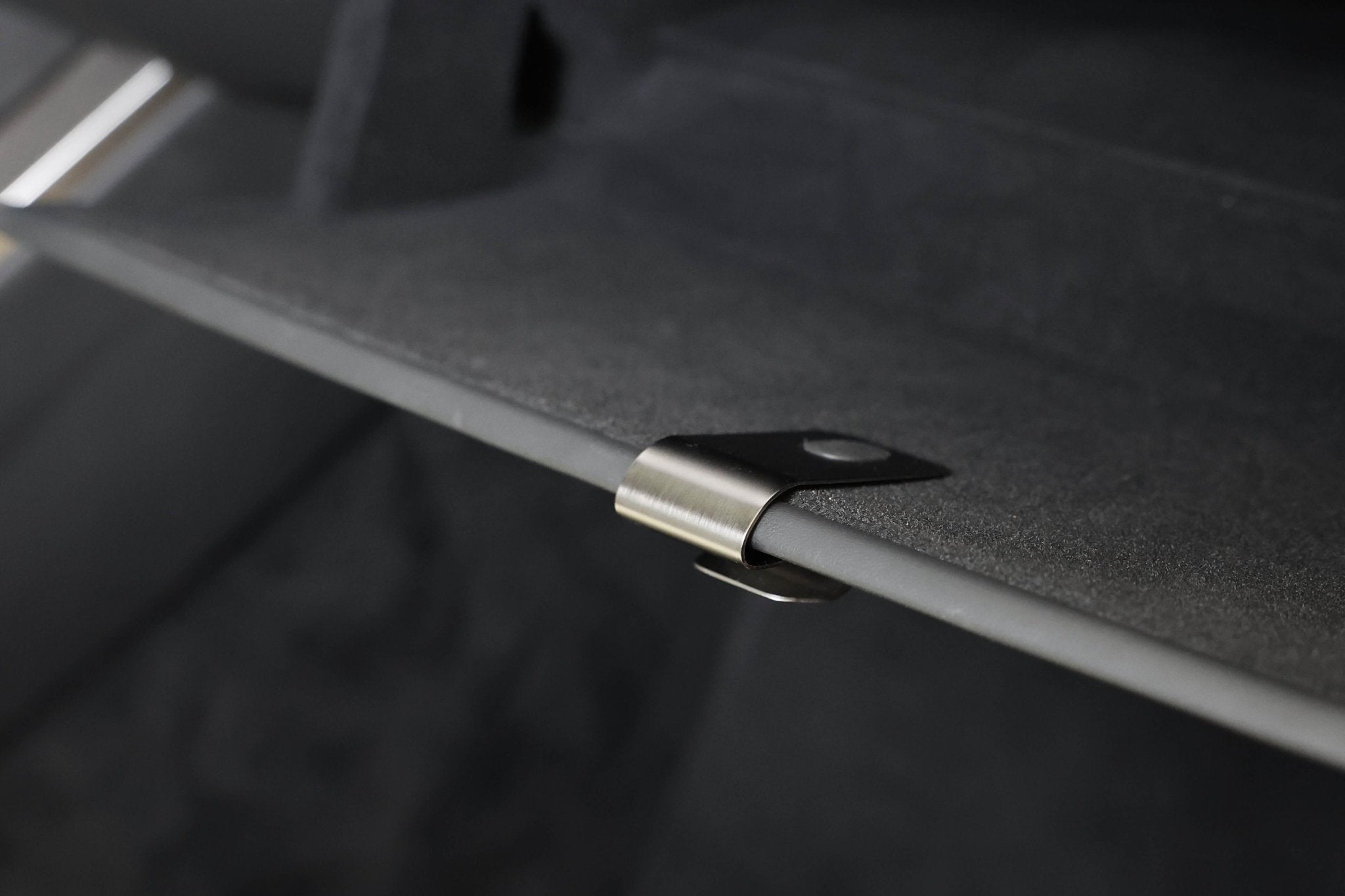 A close-up shows a stainless steel JOWUA Glovebox Hook for Tesla Model S/X/3 Highland (2024+)/Y Juniper (2025+) attached to a black, textured surface. The blurred background suggests an easy installation environment with a soft, dark ambiance.