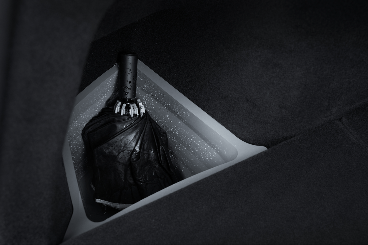 A black umbrella with water droplets rests in a triangular JOWUA Trunk Storage Bin for Model Y (2018-2024), lined with grey material and surrounded by dark fabric—likely inside a car.