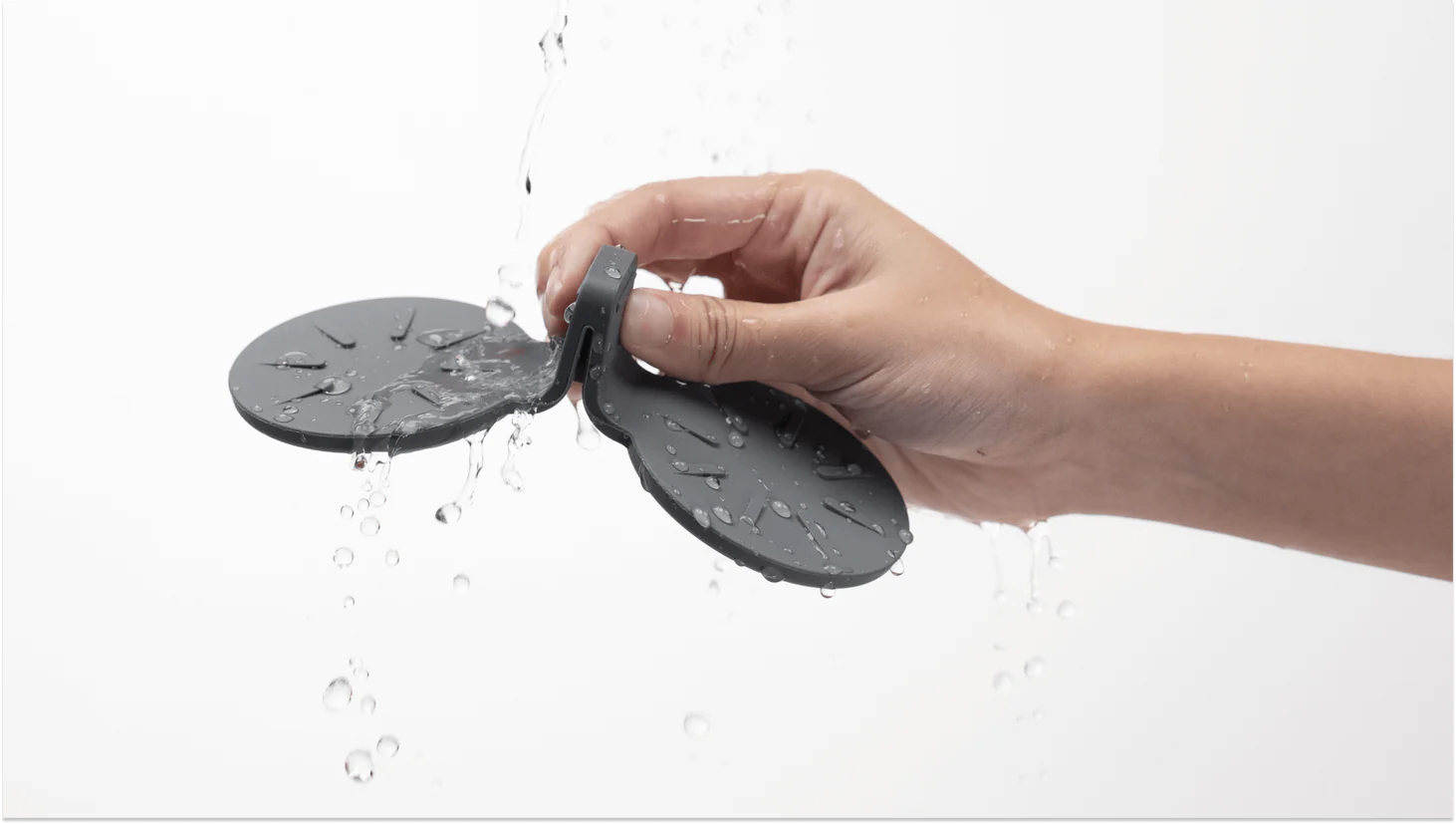 A person's hand holds a dark gray, water-resistant Rear Seat Coaster for Tesla Model S by JOWUA, crafted from premium silicone with two circular pads connected by a central piece. Water droplets cascade over the coaster and hand against a plain white background, highlighting its non-slip design.