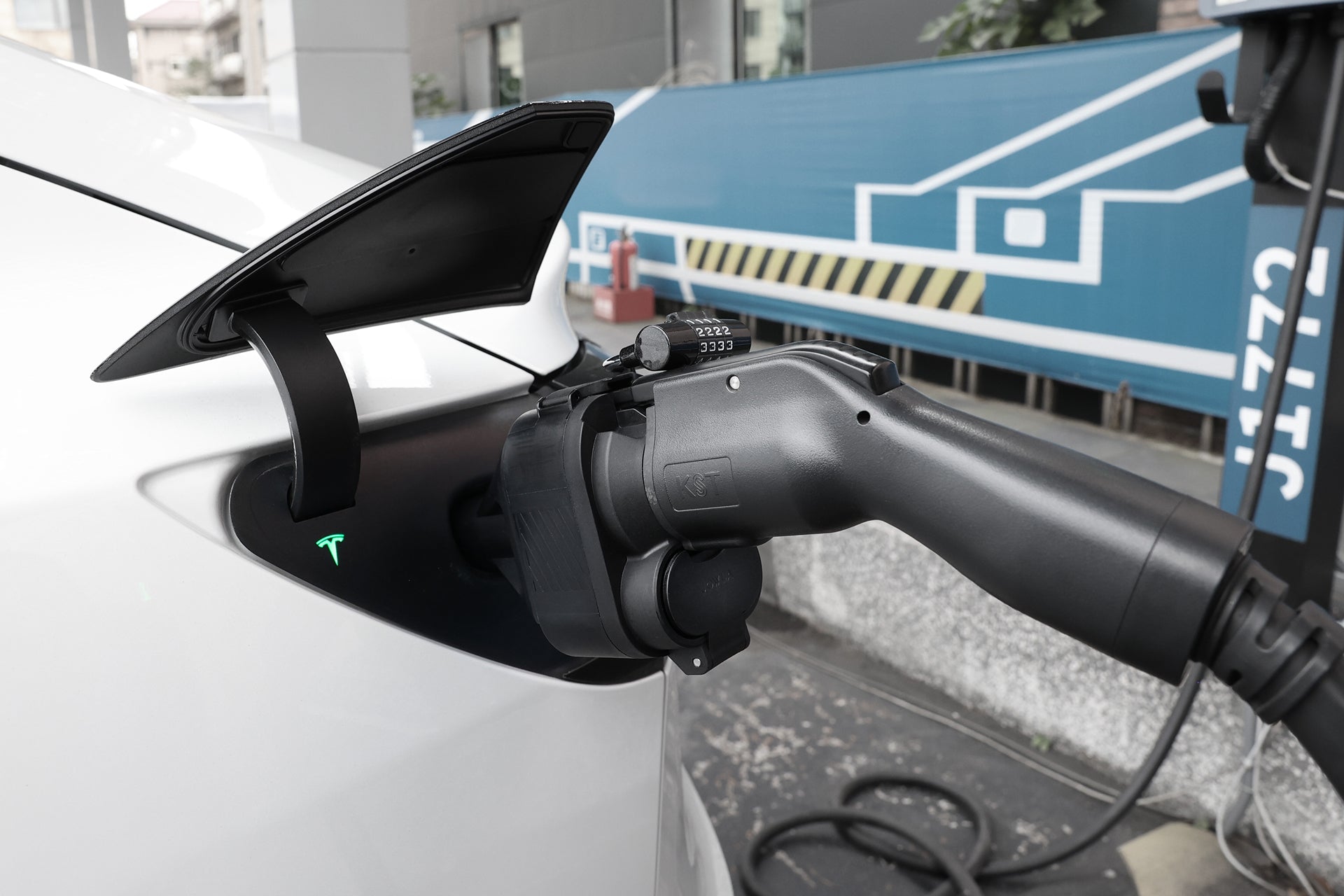 A white electric car is charged at a station using the JOWUA CCS1 to NACS DC & AC Charging Adapter for Tesla compatibility, with the cable plugged in and a blue wall in the background.