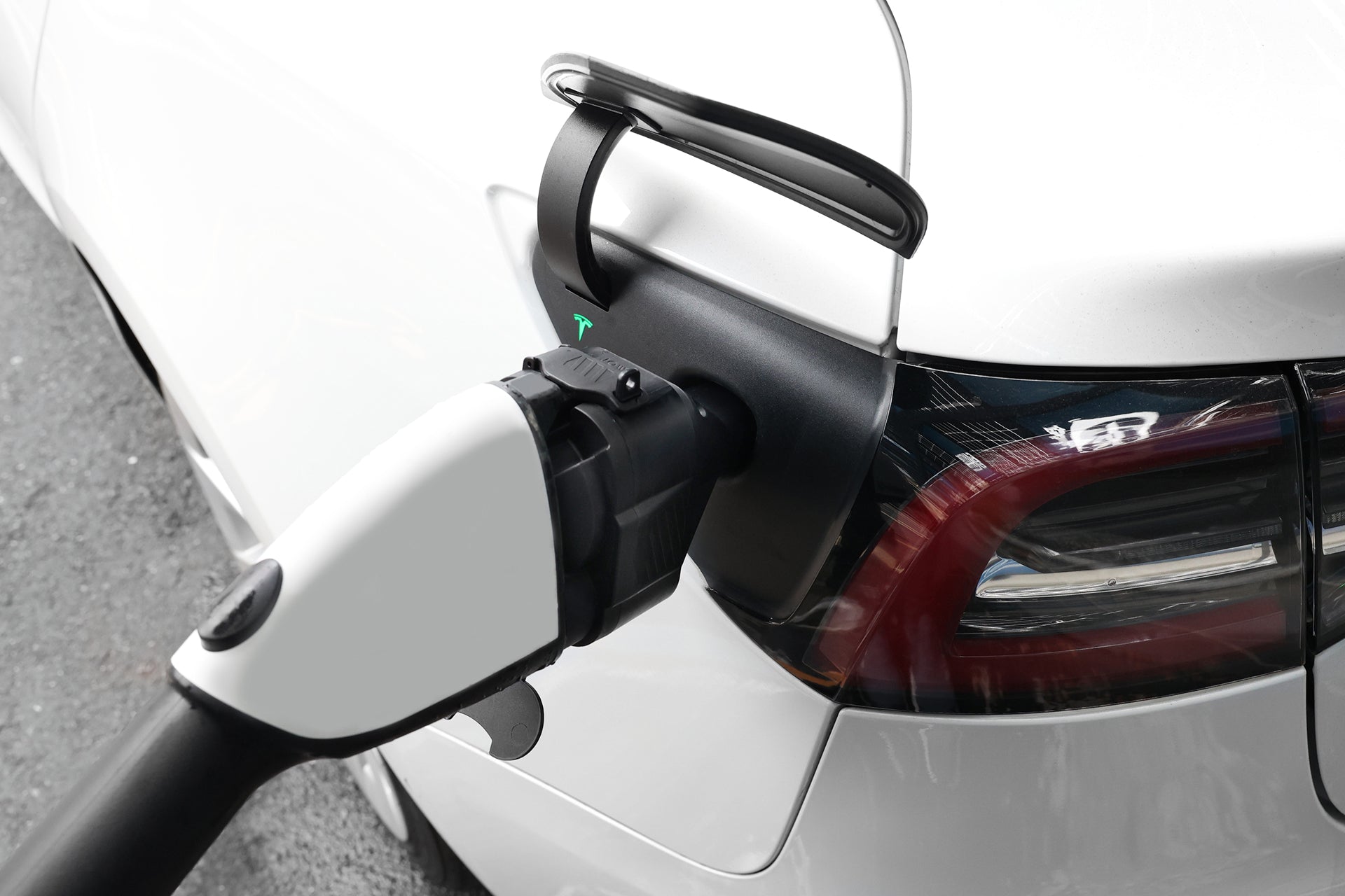 Close-up of a white car compatible with Tesla CCS using the JOWUA CCS1 to NACS DC & AC Charging Adapter, with the NACS plug connected near the rear tail light while parked on a paved surface.