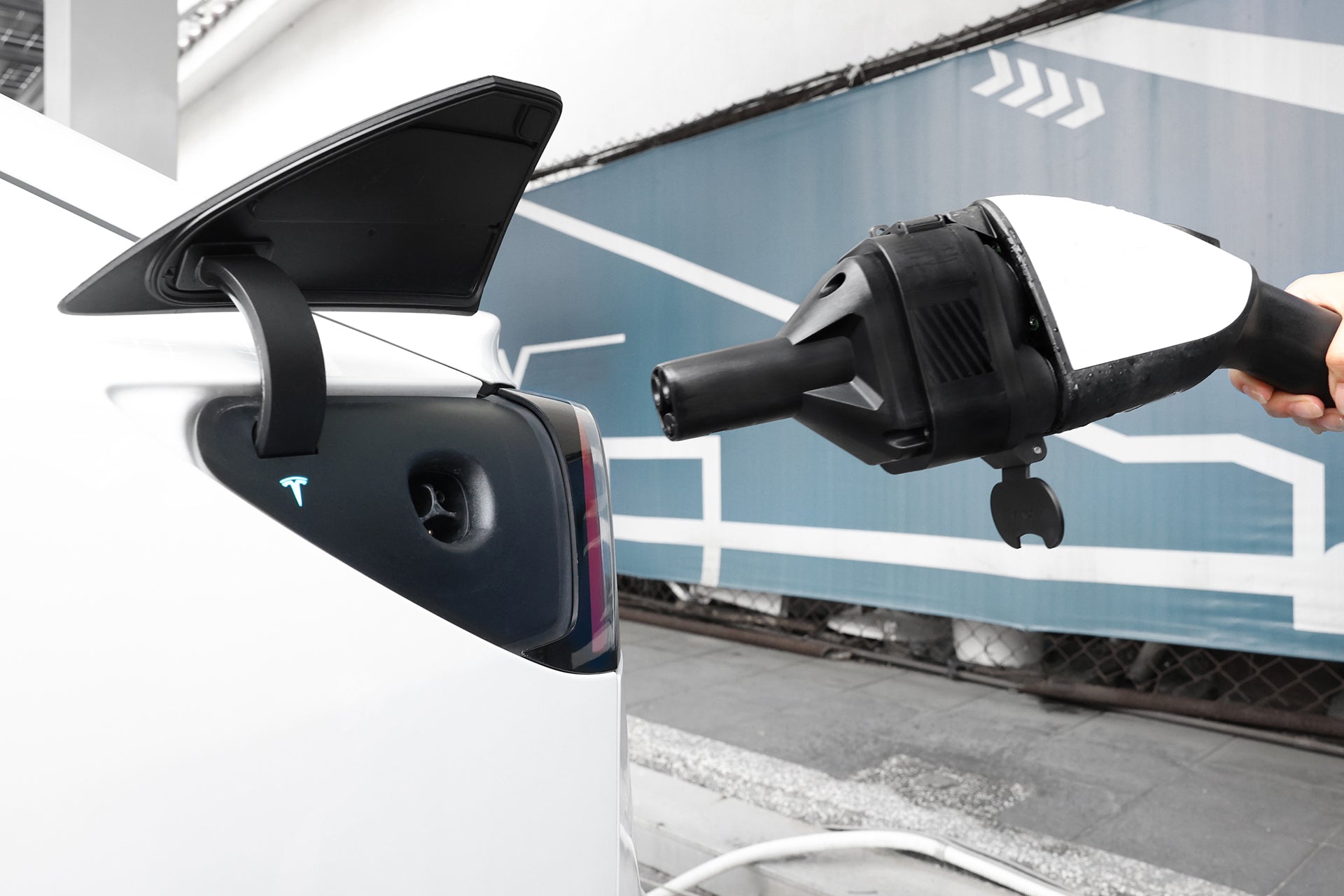 A person prepares to connect a JOWUA CCS1 to NACS DC & AC Charging Adapter to the open charging port of a white Tesla. A blue and white geometric mural is visible in the background.