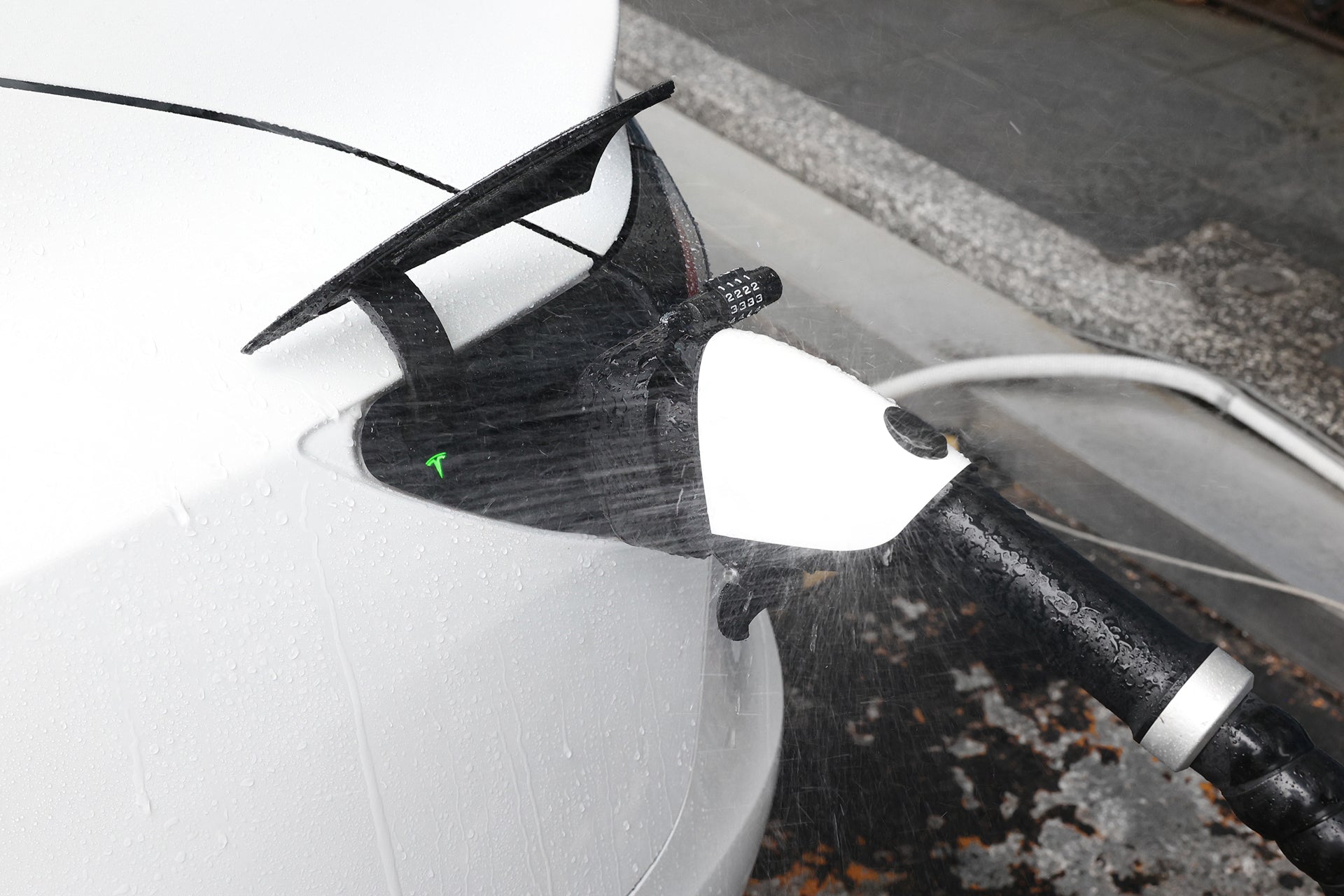 A white electric car is being charged with the JOWUA CCS1 to NACS DC & AC Charging Adapter while water sprays over the charging port and cable, demonstrating it can withstand washing during charging.