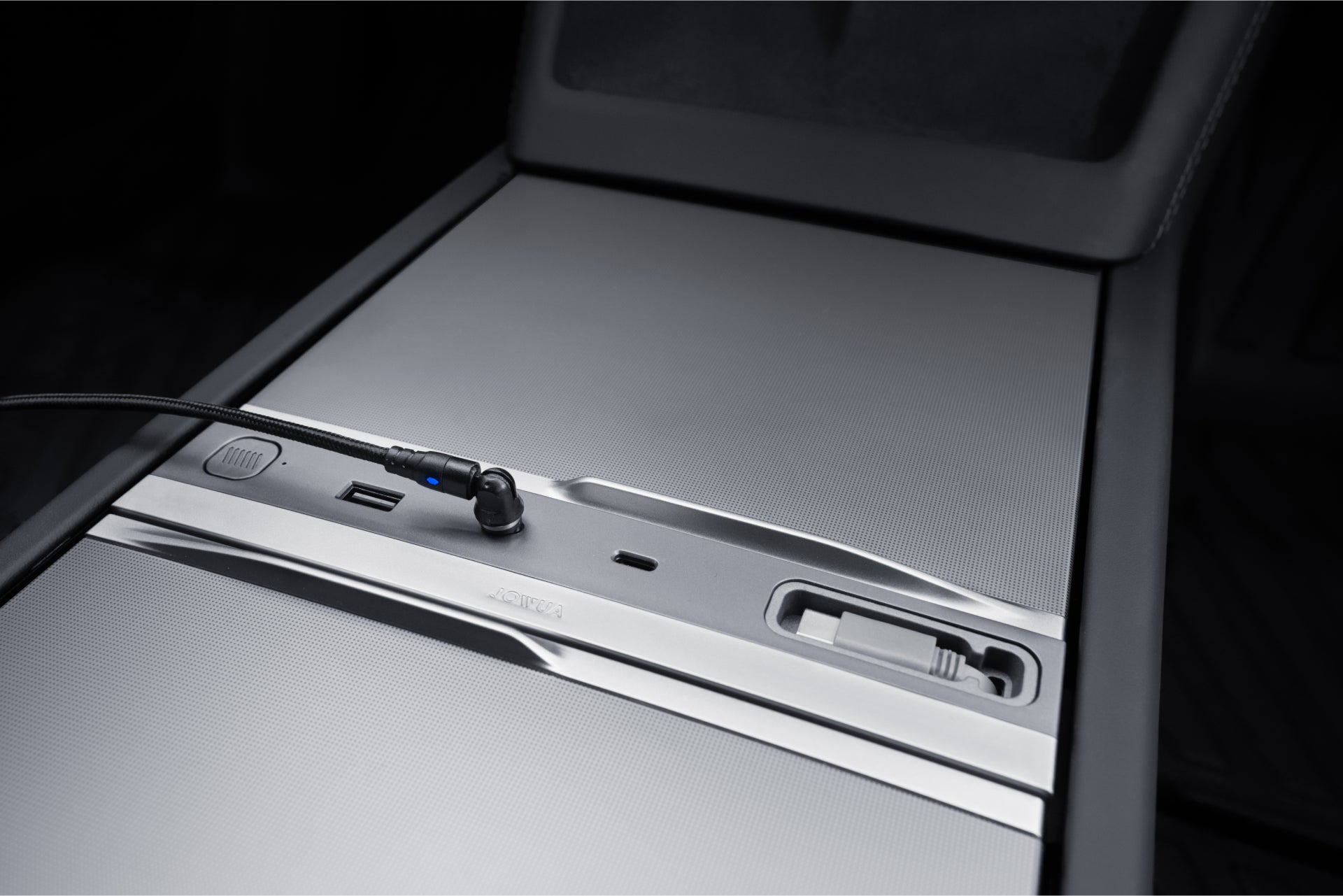A close-up of the JOWUA 130W LED Hub for Tesla Model Y Juniper 2025+ / Model 3 2024+ shows a sleek metallic surface with a USB port, audio jack with cable, and recessed slider with opening, offering minimalistic and modern charging and storage.