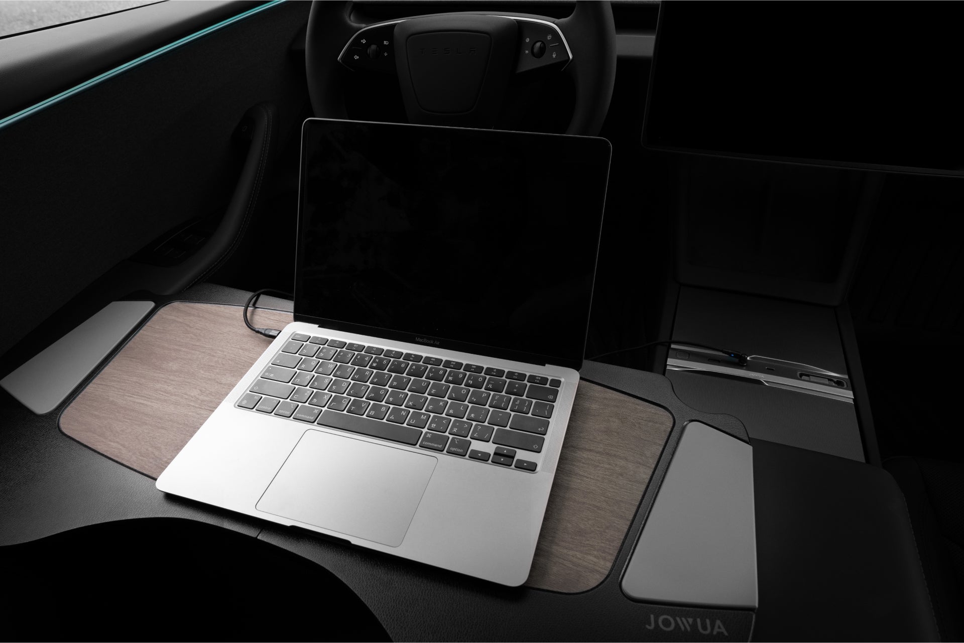 A laptop with a partially closed lid rests seamlessly on the Tesla center console's dashboard tray, blending with the minimalist interior and steering wheel, powered by JOWUA's 130W LED Hub for Tesla Model Y Juniper 2025+ / Model 3 2024+.